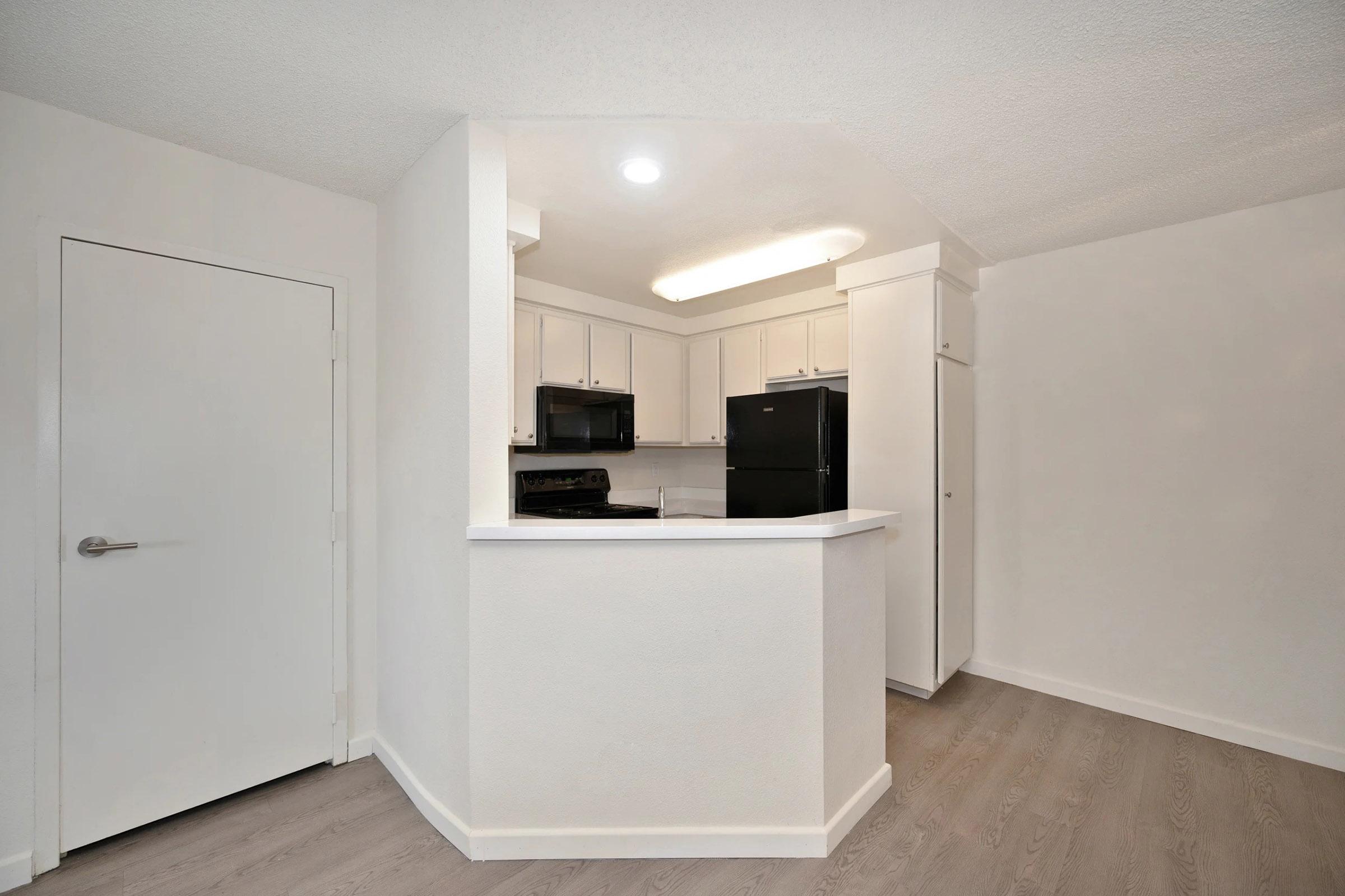 A modern kitchen featuring an open layout with white cabinetry, black appliances, and a light-colored countertop. The space has light wood flooring and a bright, clean aesthetic, perfect for a contemporary living environment.