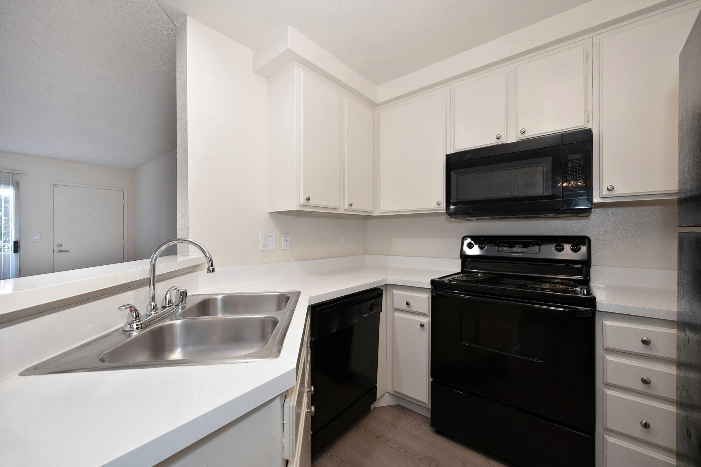 Modern kitchen featuring white cabinetry, a double sink, a black microwave, and an oven. The countertop is light, complementing the appliances. There's a dishwasher next to the sink, and the floor is light-colored wood. A doorway in the background leads to another room.