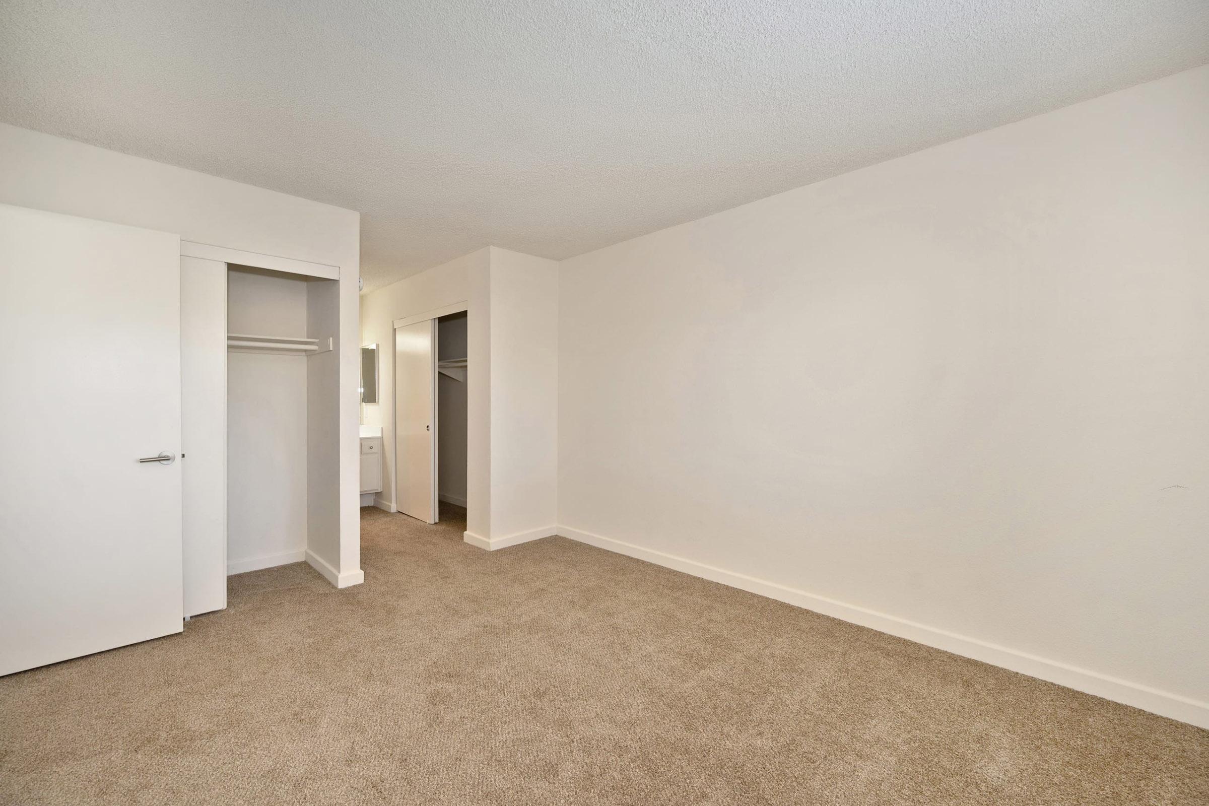 A spacious, empty room with light beige carpet and white walls. There are two closet doors on the left, and a doorway leading to another area. The space is well-lit and has a neutral, minimalist design, making it suitable for various uses or decorations.
