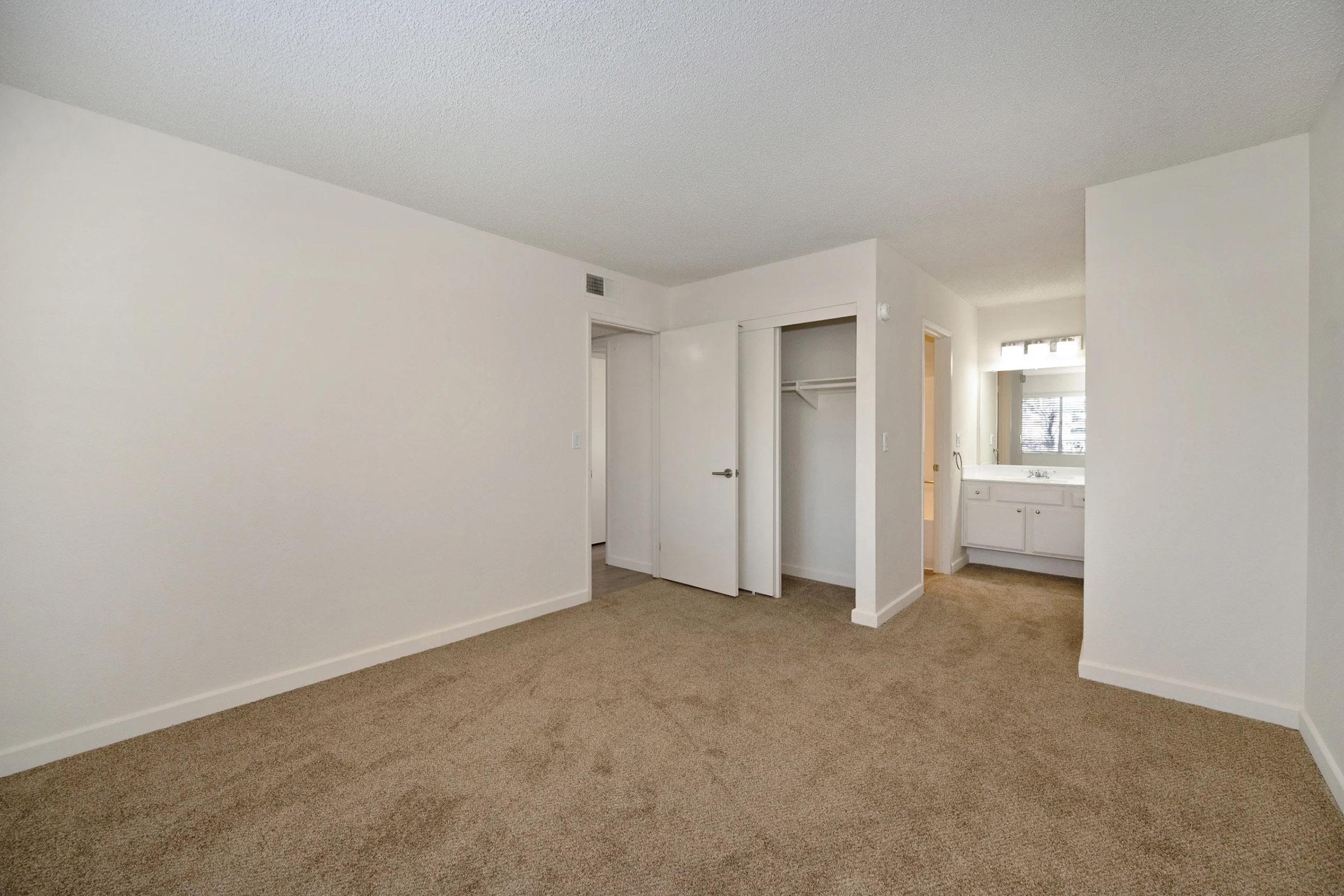 A vacant room with beige carpet and light-colored walls. There are two doors leading to an adjoining room and a closet. A bathroom area with a vanity is visible in the background. Natural light enters through a window, creating a bright and open atmosphere.