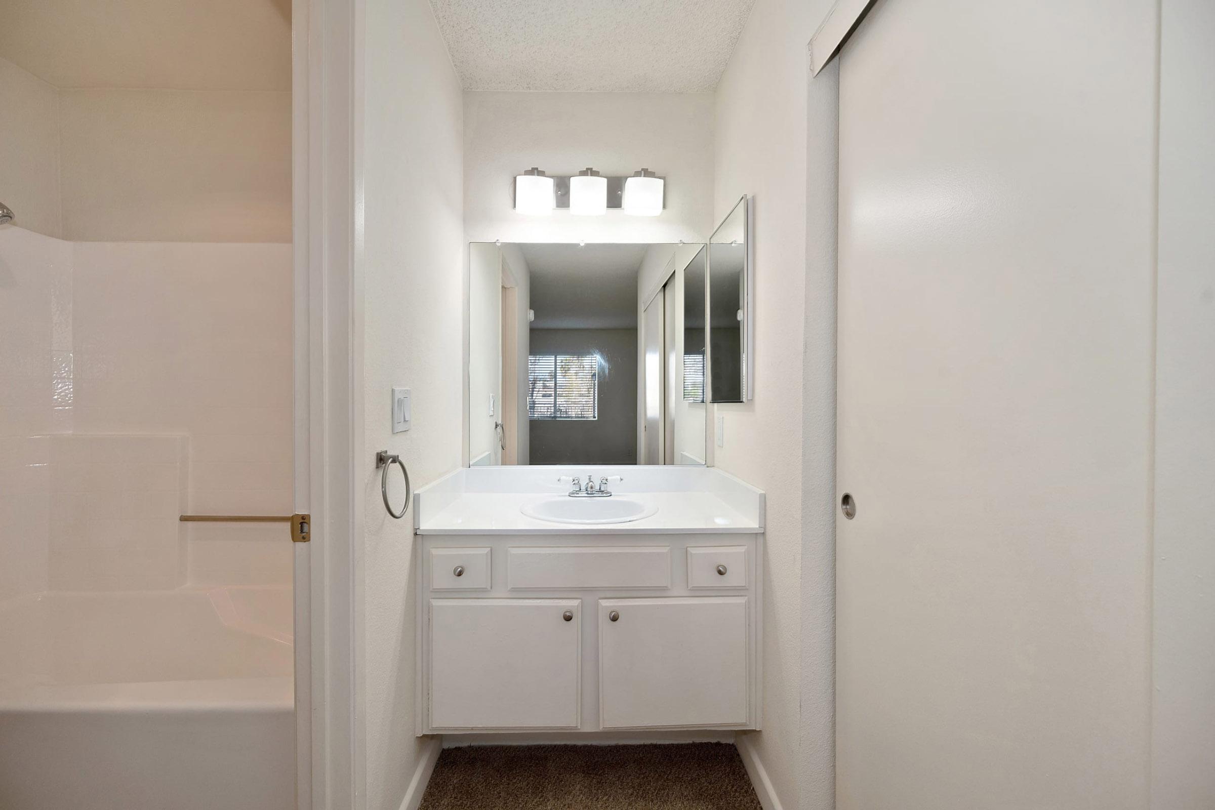 A clean and simple bathroom featuring a white vanity with a sink and mirror. There is a shower and bathtub in the background, and a door leading to another room. The walls are painted in neutral tones, and the lighting is provided by a modern fixture above the mirror.