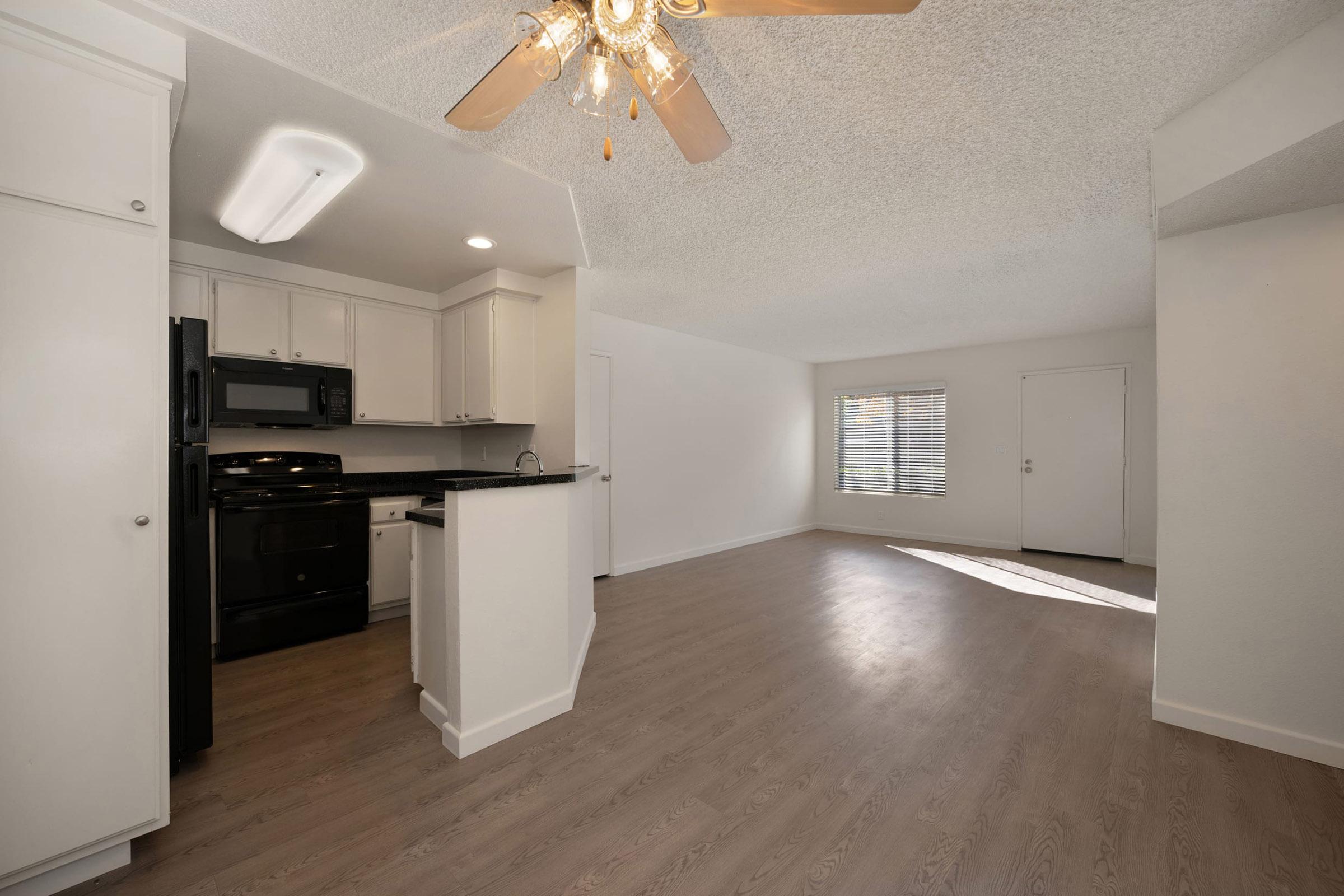 Modern kitchen and living area in an apartment. The kitchen features white cabinets, dark countertops, and black appliances. A ceiling fan hangs above, and large windows provide natural light into the spacious, open living room with a light-colored floor and white walls.