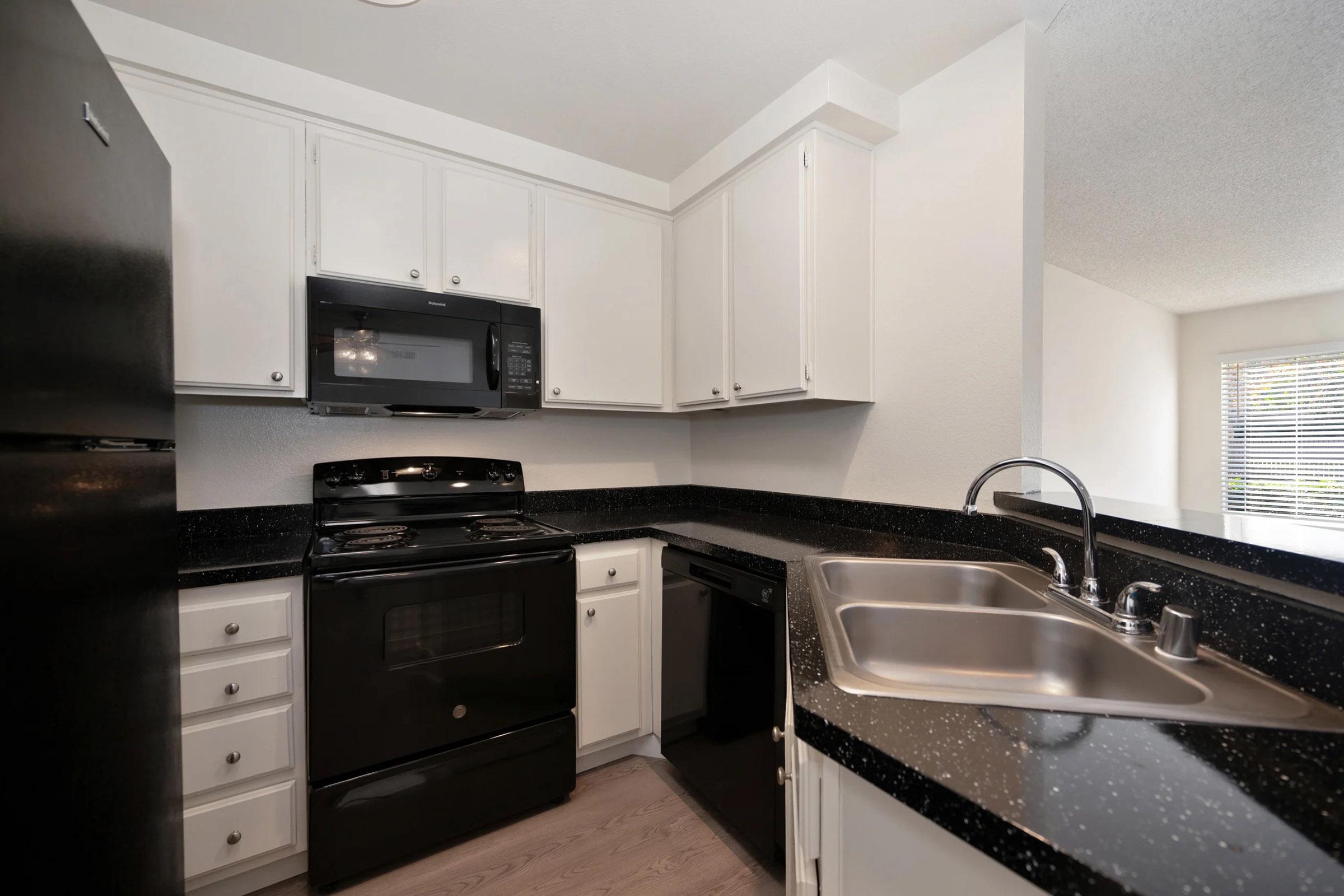A modern kitchen featuring black appliances, including a refrigerator, microwave, and oven. White cabinets line the walls, complemented by a dark granite countertop. A double sink is visible, and there is a window that brings in natural light. The flooring is light-colored wood, enhancing the space's contemporary look.