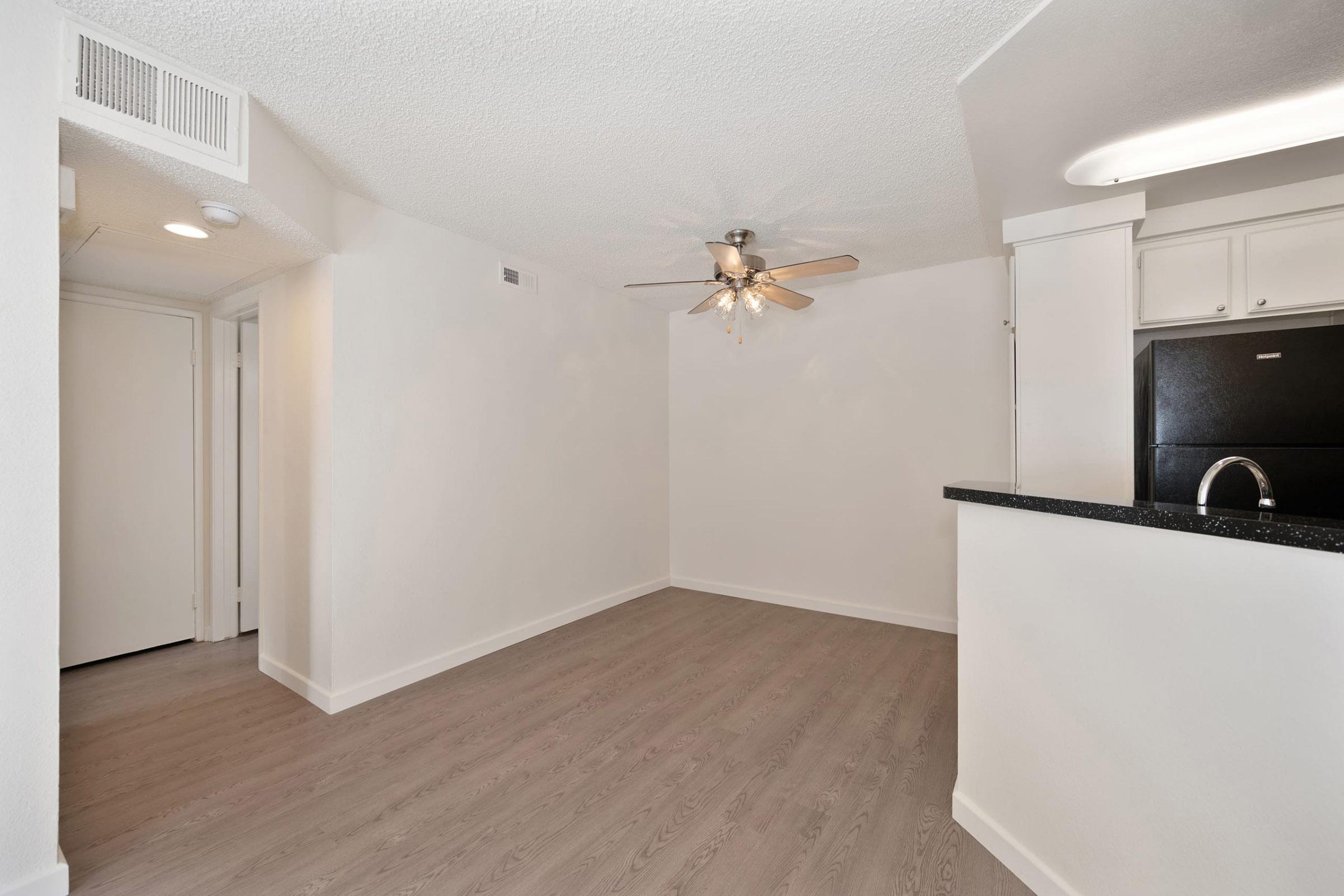 A bright, empty room featuring light-colored walls, a ceiling fan with wooden blades, and wood-like flooring. The space has an open layout connecting to a kitchen with black appliances and a granite countertop. Natural light may enter from an unseen window.