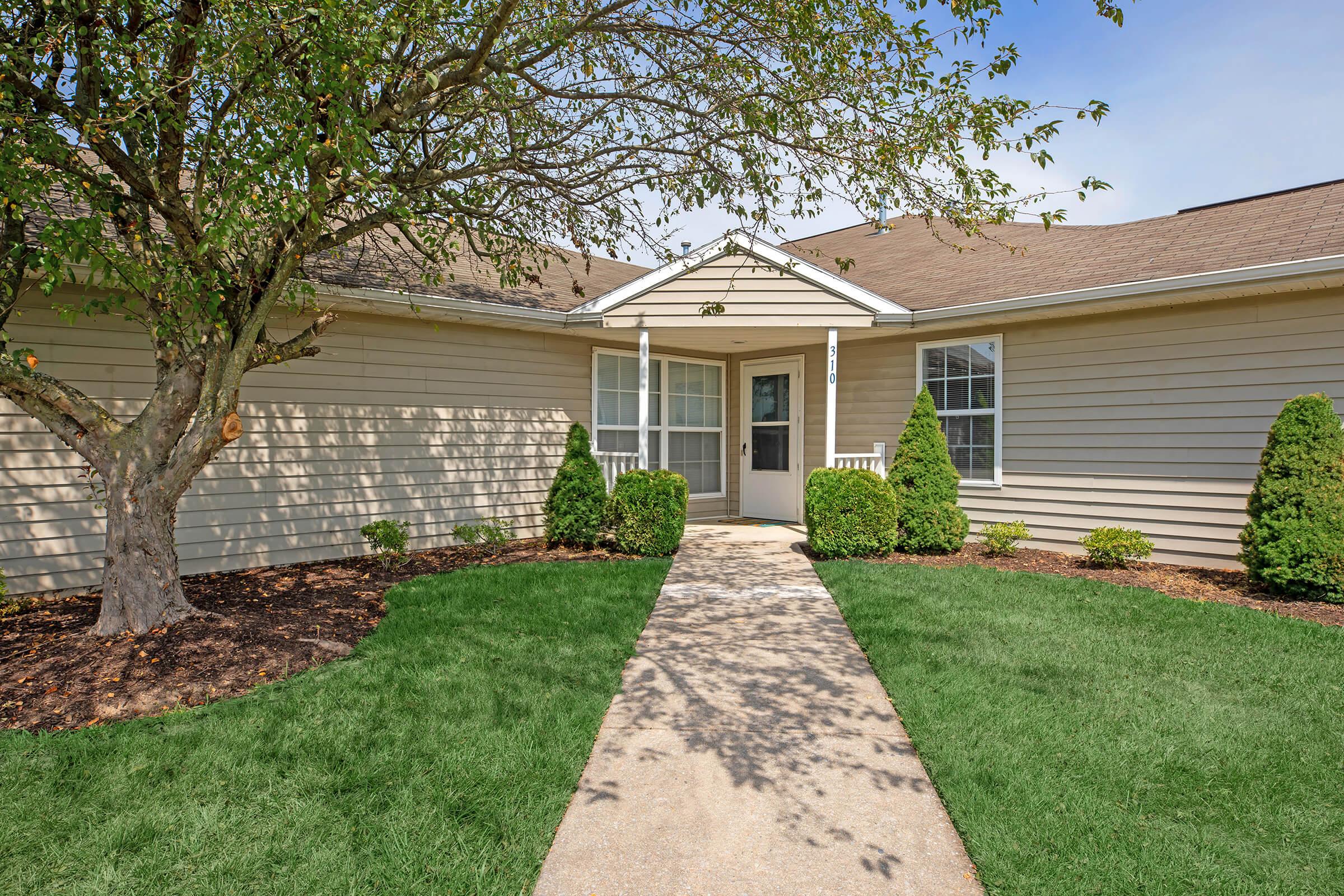 A welcoming entrance to a light-colored single-story home surrounded by well-maintained greenery. The pathway leads to a front door, framed by two neatly trimmed shrubs and a small tree nearby. A clear blue sky complements the inviting atmosphere of the property.