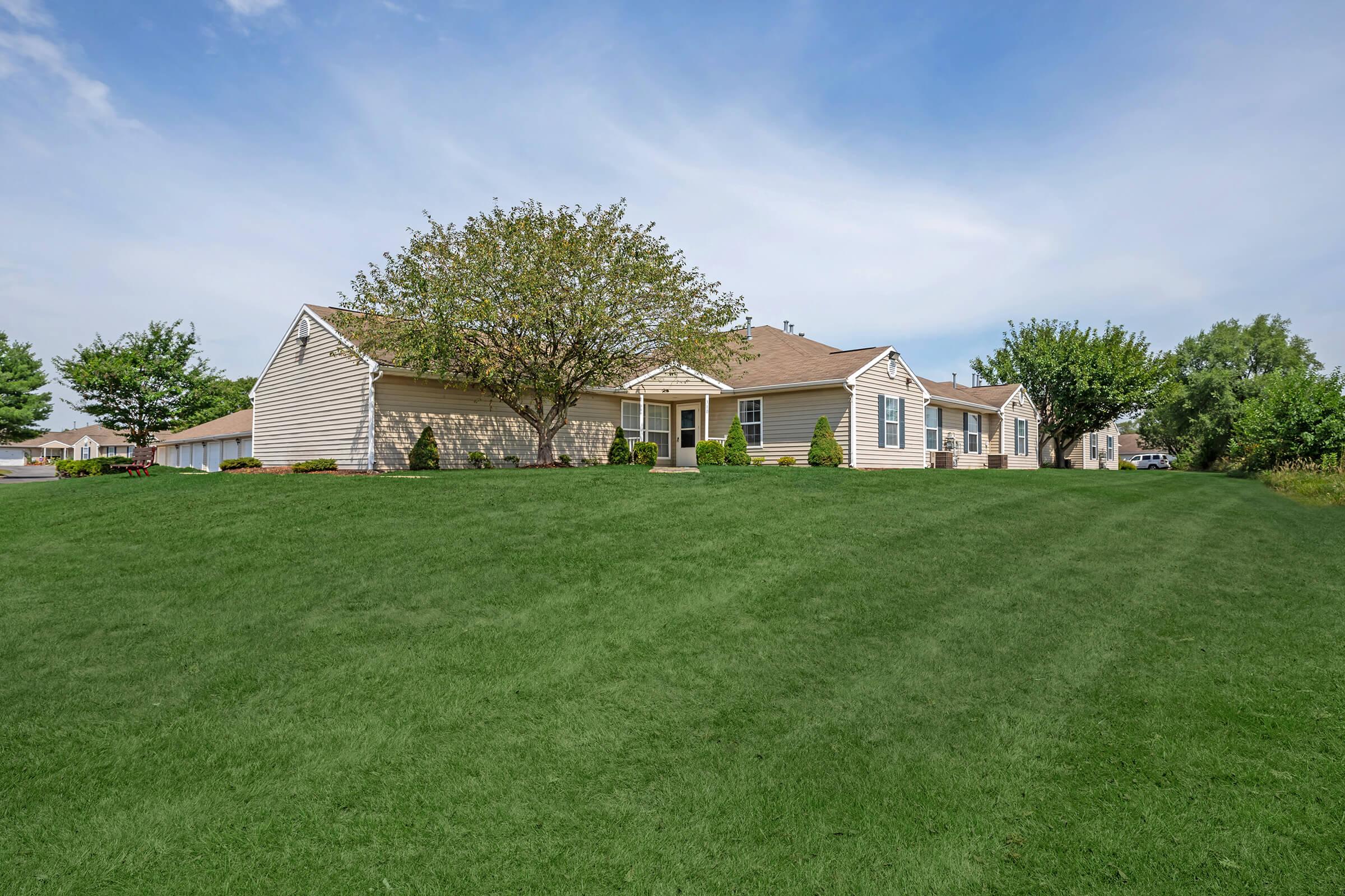 A well-maintained single-story house surrounded by lush green grass and neatly trimmed shrubs. The home features a light-colored exterior with multiple windows and a sloped roof. A tree stands prominently in the front yard, offering shade. The setting is serene and inviting, with a clear blue sky overhead.