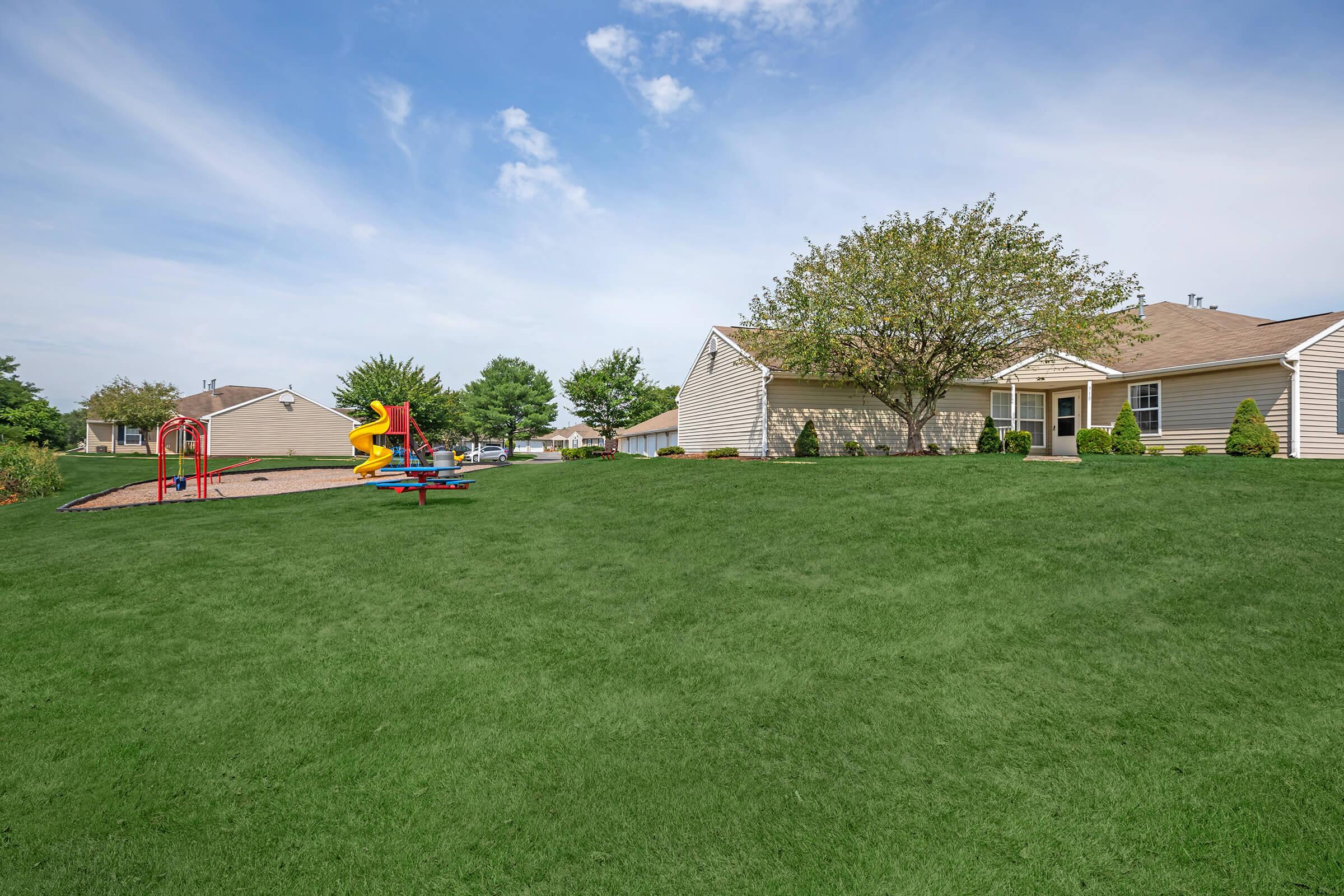 A well-maintained playground with colorful equipment, including a slide and climbing structure, set on a lush green lawn. In the background, there are residential buildings with landscaped surroundings under a clear blue sky.