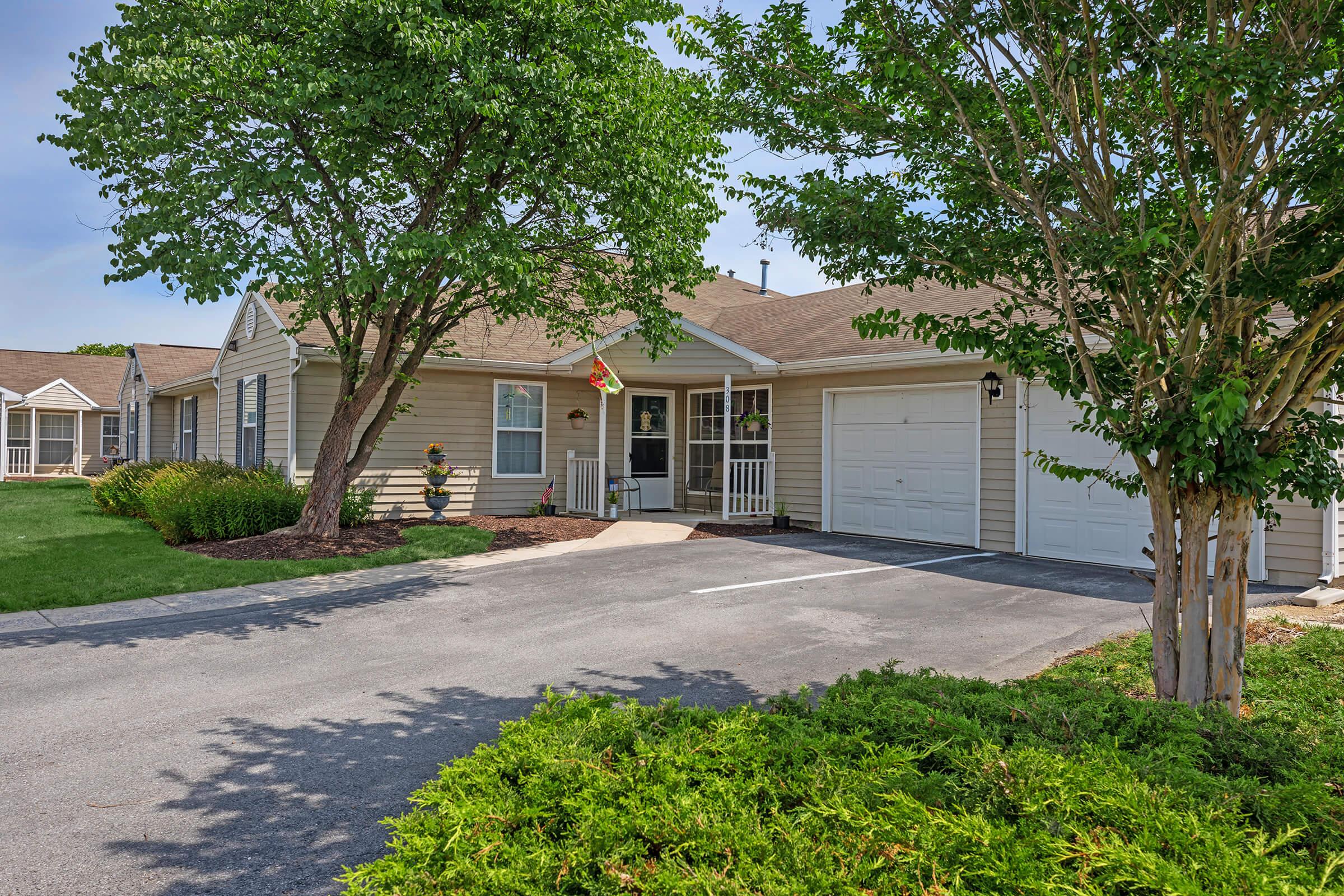 A single-story residential home with a well-maintained front yard. The house features beige siding, a covered entryway with a decorative flag, and a one-car garage. Trees and shrubs enhance the landscaping, with a paved driveway leading to the garage and front entrance.