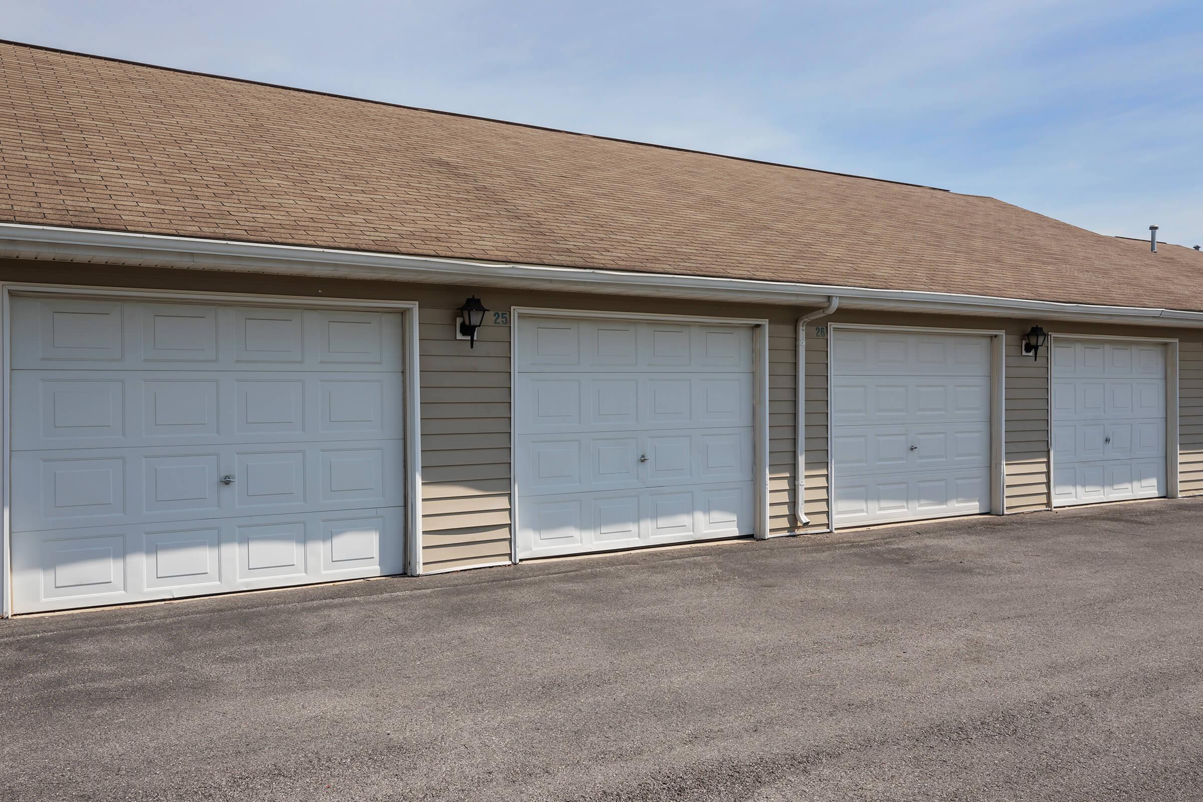 Row of three white garage doors with rectangular panels, set against a light tan, vertical siding exterior and a brown shingled roof. The driveway is paved and leads up to the garages, with clear blue sky above. Two lantern-style lights are visible beside the doors.