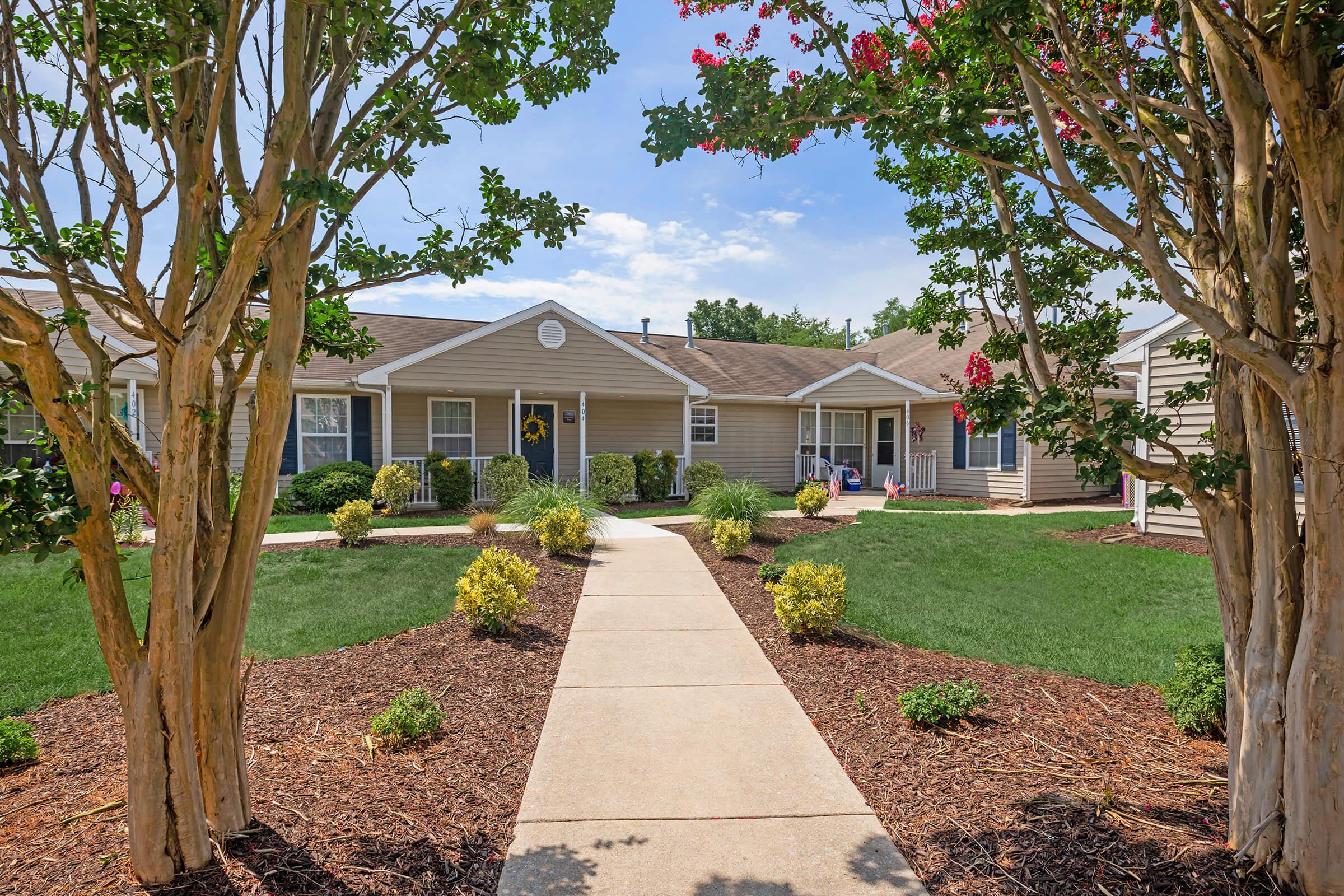 A well-maintained pathway leading through lush green grass and landscaped gardens, framed by flowering trees. Small shrubs and flower beds line the sides, with single-story homes visible in the background under a clear blue sky. The scene exudes a welcoming and peaceful residential atmosphere.