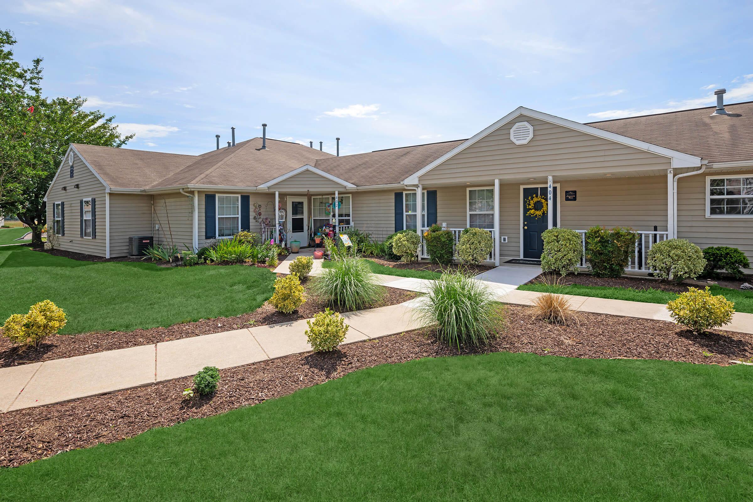 A well-maintained single-story apartment building with light-colored siding and a welcoming front porch. Lush green lawns and landscaped flower beds featuring bushes and small plants line the path leading to the entrance, with a bright blue sky overhead.