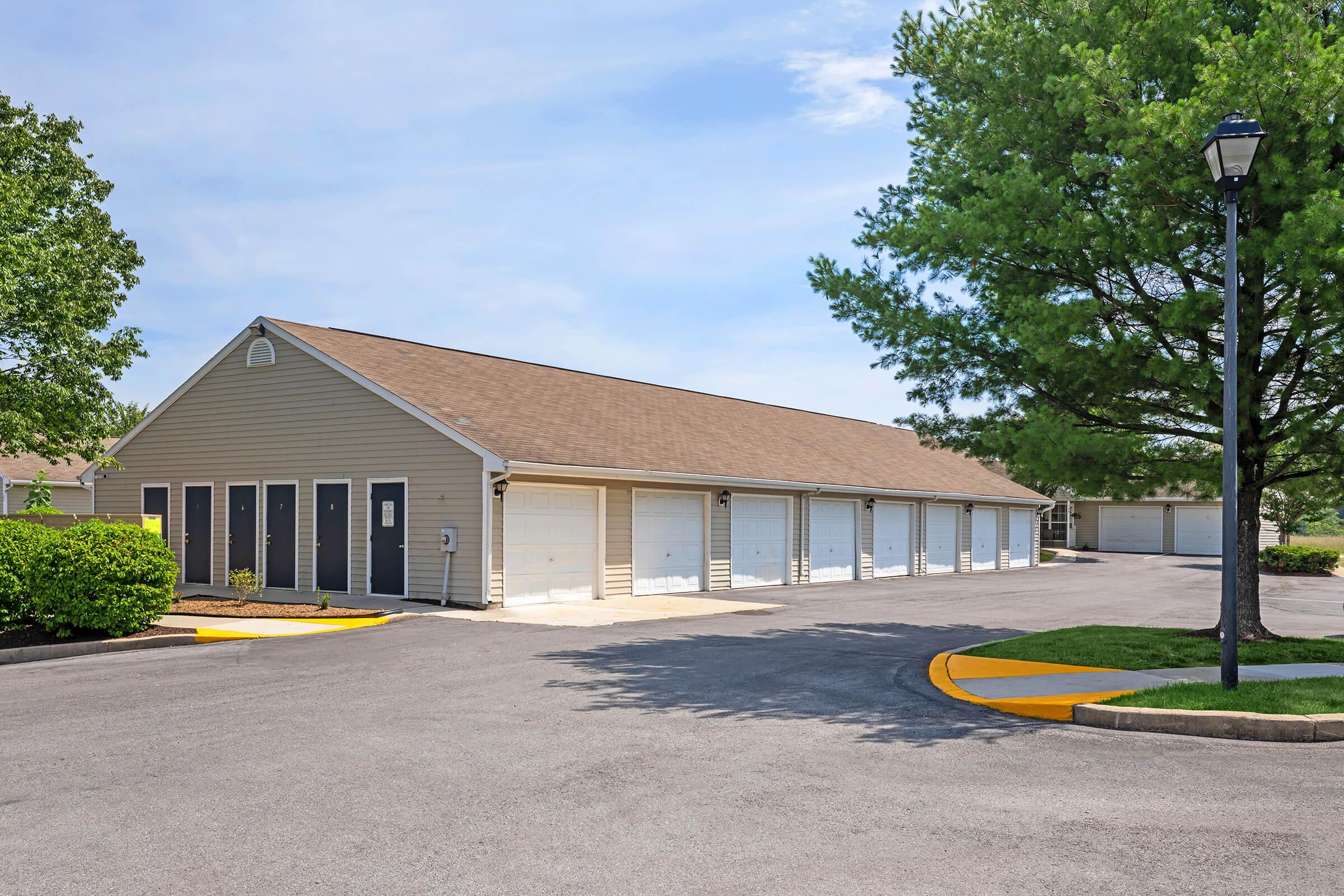 A well-maintained storage facility featuring multiple garage doors arranged in a single row. The building has a light-colored exterior with a brown roof. Surrounding landscaping includes green bushes and trees, under a clear blue sky. A lamp post is visible along the pathway.