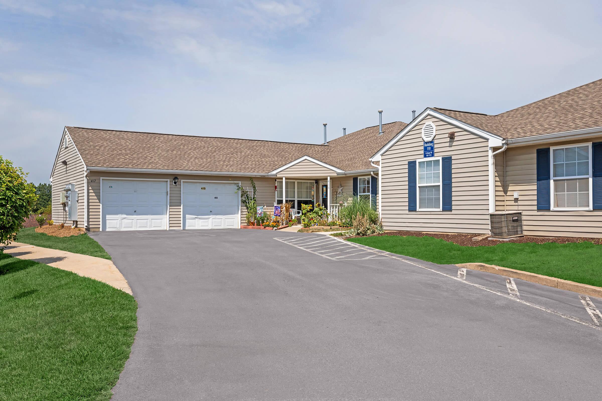 A single-story residential building with a beige exterior and a brown roof. The front features a driveway leading to a garage, lush green landscaping, and a small porch area. Windows are framed with blue shutters, and the entrance is adorned with flower pots and plants. The sky is clear, indicating a sunny day.