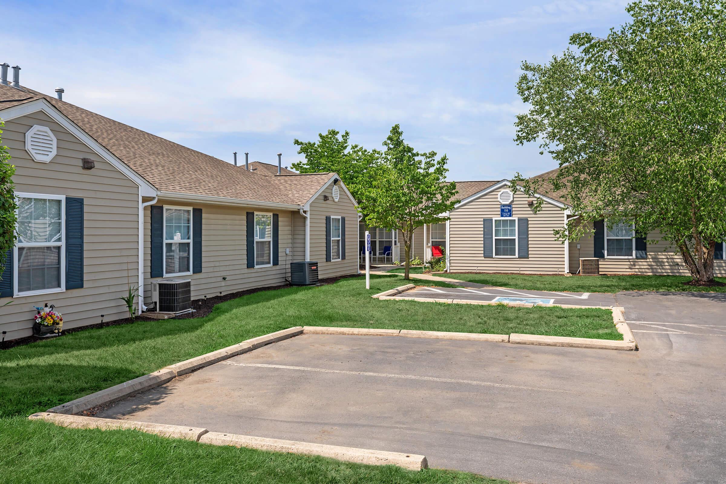 A picturesque view of a residential complex featuring two single-story houses with beige siding and blue shutters. Green grass surrounds the buildings, and small trees provide shade. A paved parking area is visible in the foreground, with a clear blue sky above.