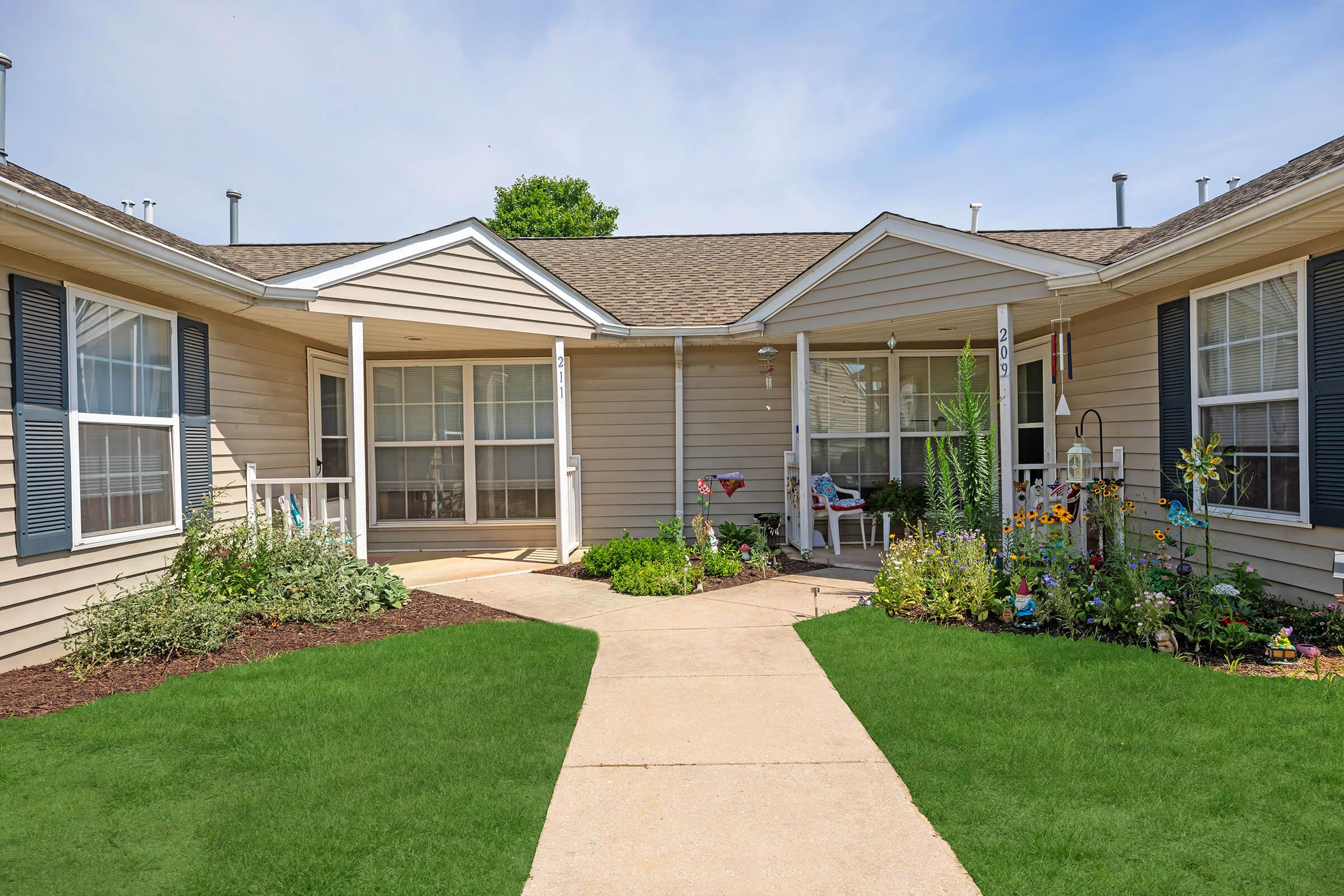 A well-maintained courtyard featuring a paved walkway surrounded by green grass and colorful flower gardens. The building has beige siding, multiple windows with white trim, and decorative outdoor furniture. Bright plants and garden decorations add charm to the inviting outdoor space.