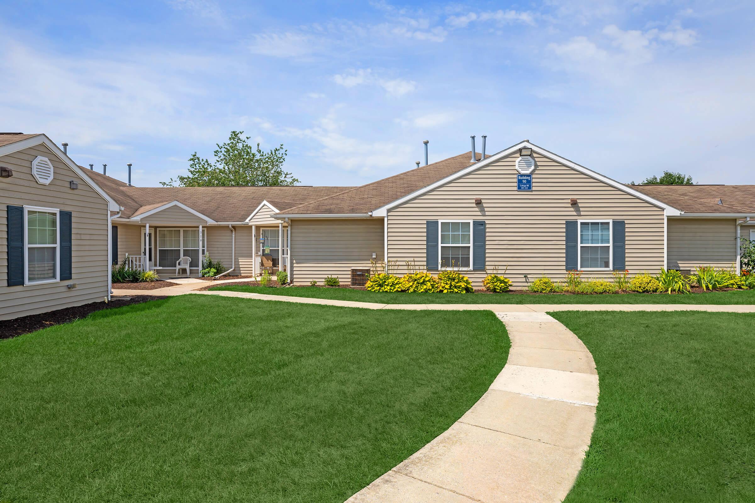 A well-maintained single-story building with beige siding and blue accents, featuring a landscaped garden with yellow flowers and a green lawn. A paved pathway leads to the entrance, with additional structures visible in the background under a clear blue sky.