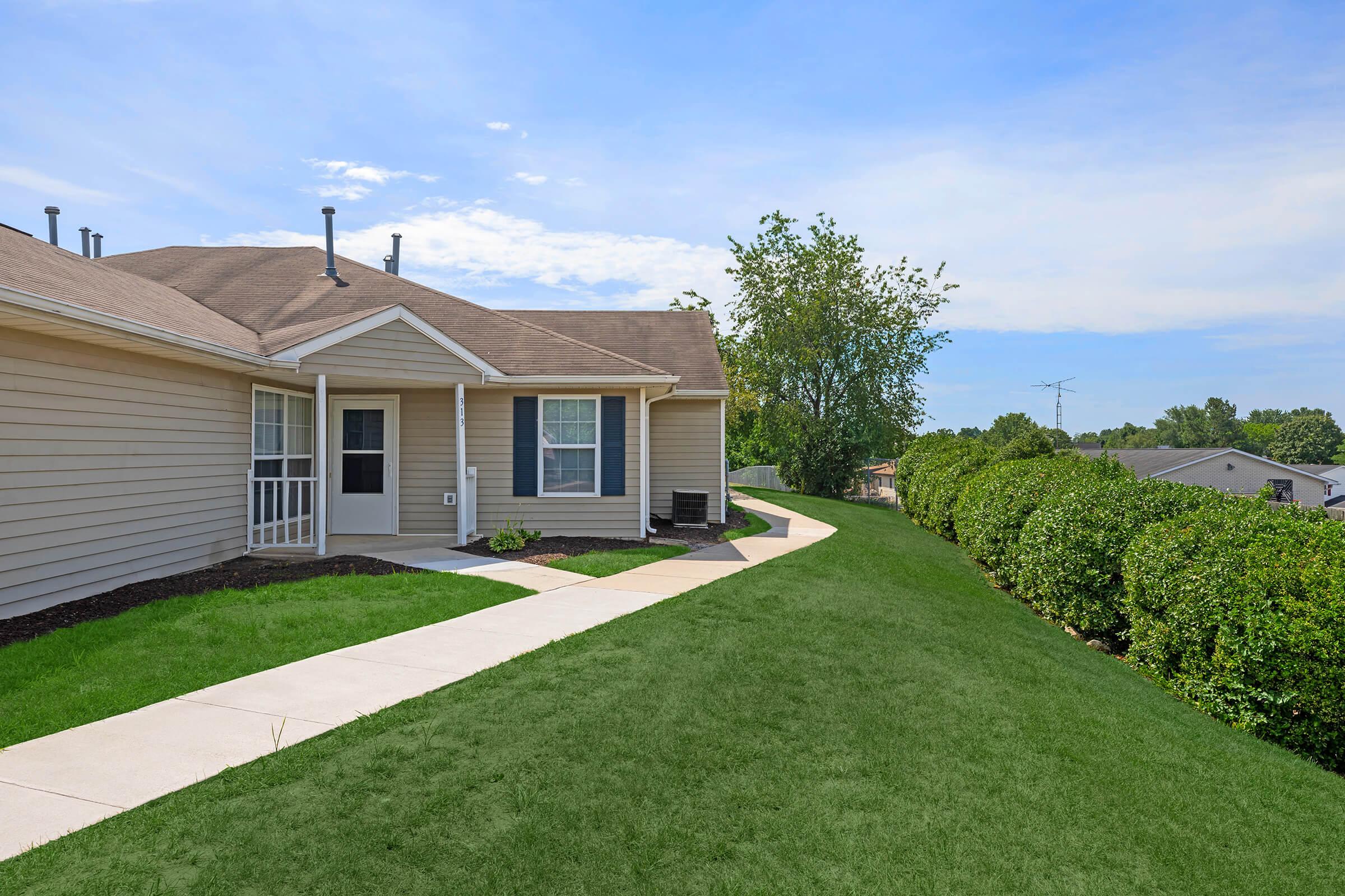 A well-maintained single-story house with a light beige exterior, featuring a small porch and large windows. A neatly paved pathway runs along a lush green lawn, leading to a manicured hedge on one side. The sky is clear with some clouds, adding a bright atmosphere to the scene.