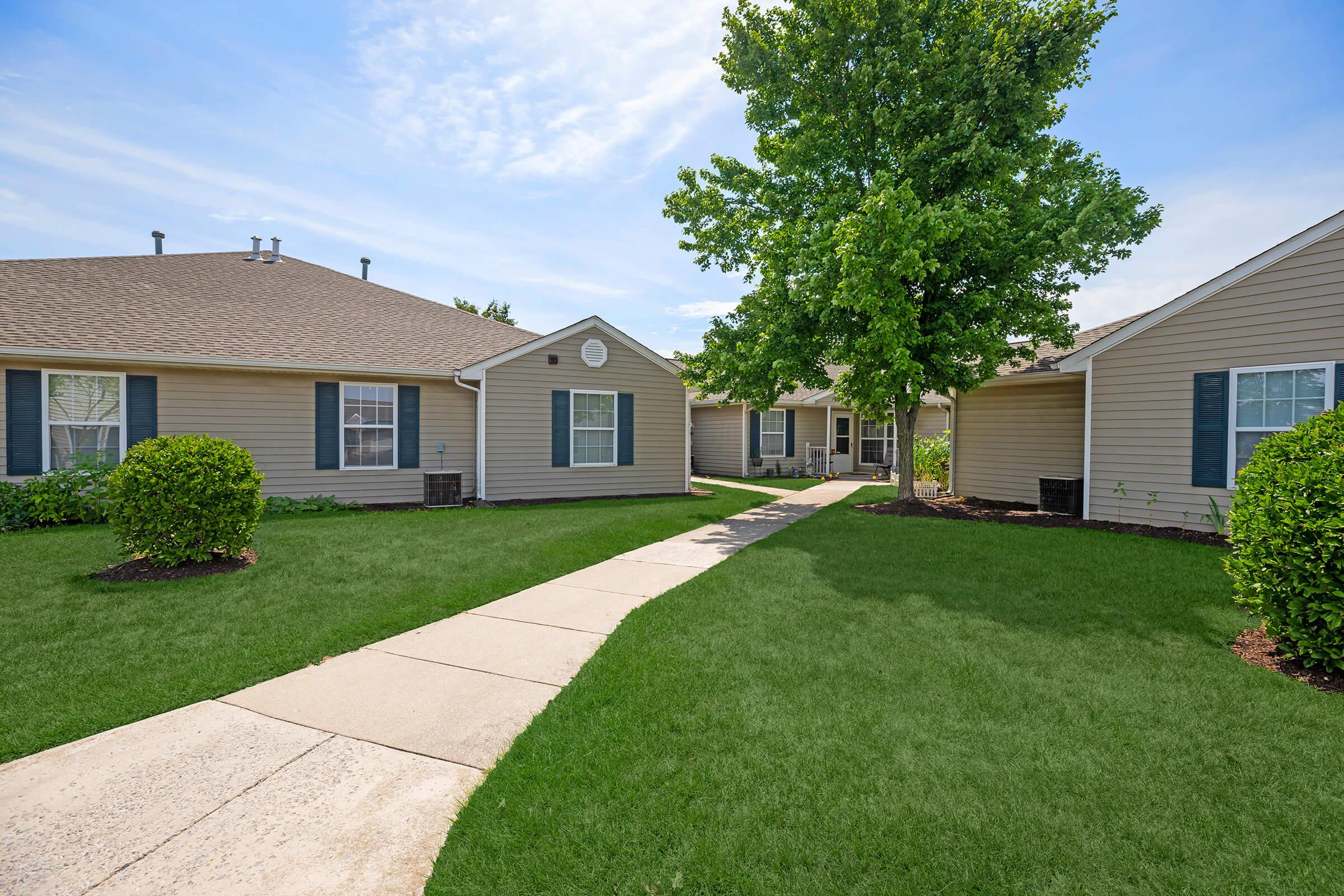 A well-maintained residential complex featuring beige single-story buildings with green shutters. A paved walkway leads through a manicured lawn with shrubs and a large tree, under a clear blue sky. The environment appears inviting and peaceful.