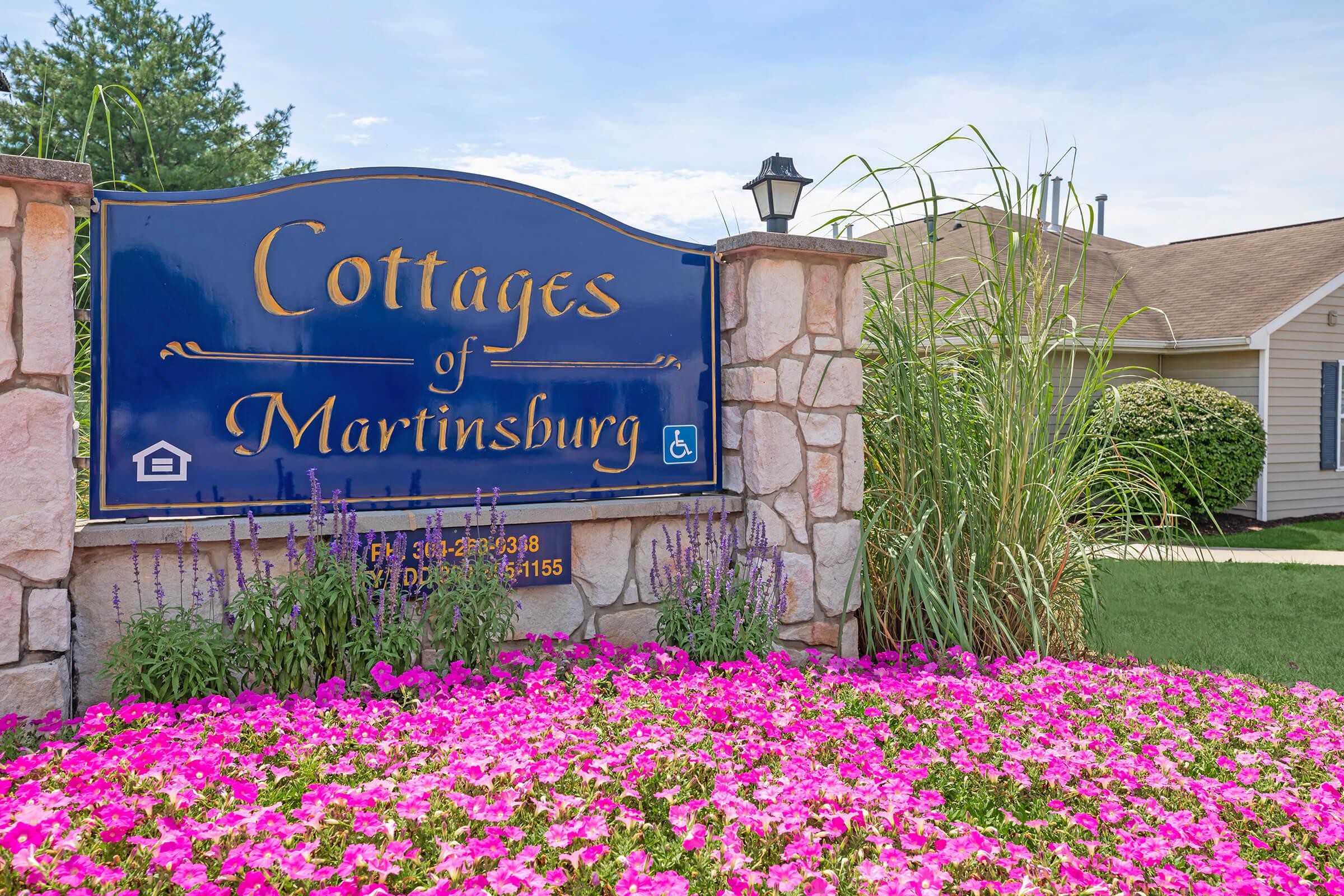 Sign for "Cottages of Martinsburg" featuring a blue background with white text. Surrounded by colorful flowering plants, primarily pink petunias. A lamppost is visible near the sign, and there is a clear blue sky in the background. The location appears cozy and welcoming.
