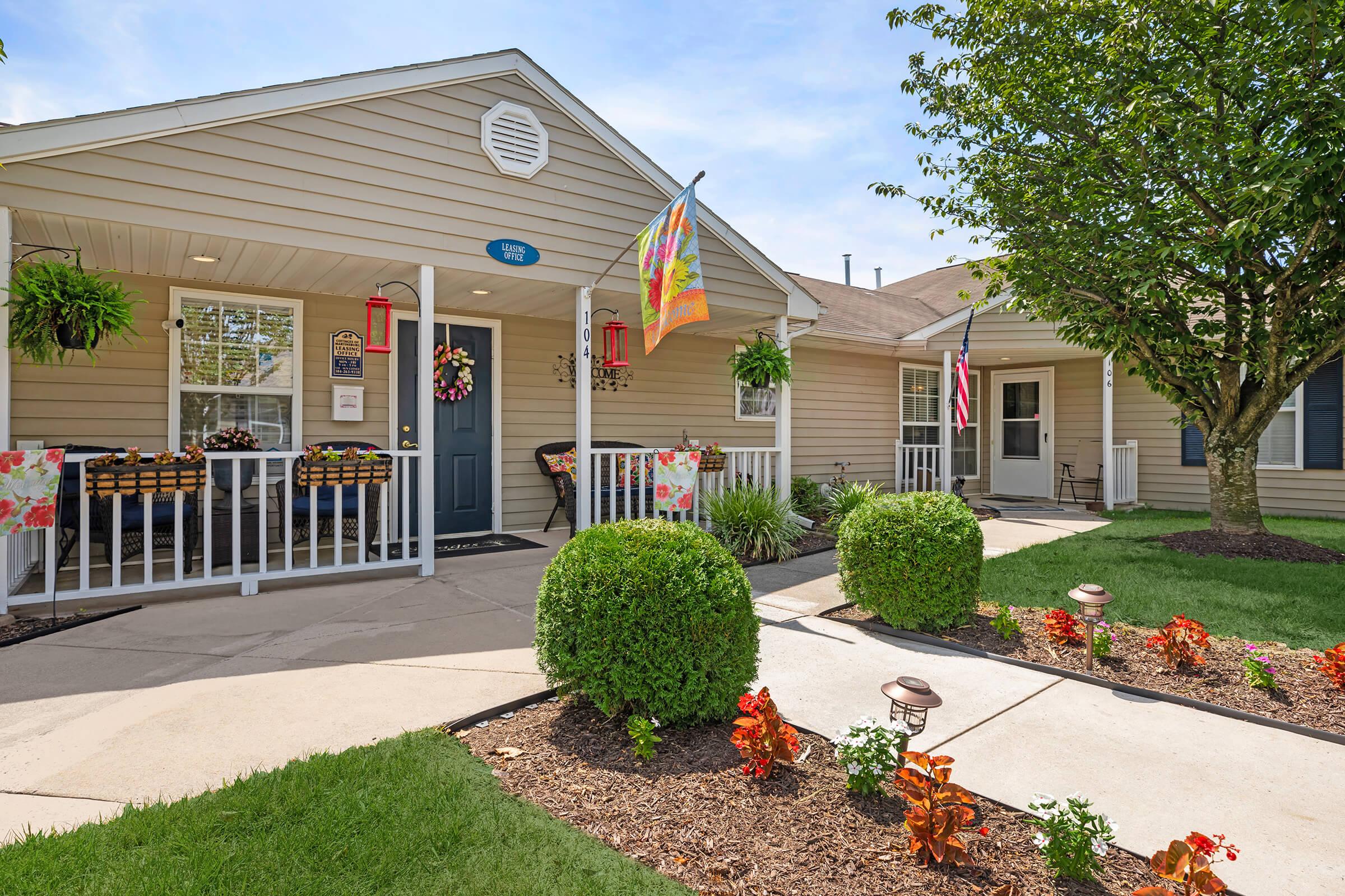 A charming single-story home featuring a light-colored exterior with a welcoming front porch adorned with decorative plants and a festive flag. A neat lawn with colorful flower beds and a paved pathway leads to the entrance. The porch has seating areas, creating an inviting atmosphere.