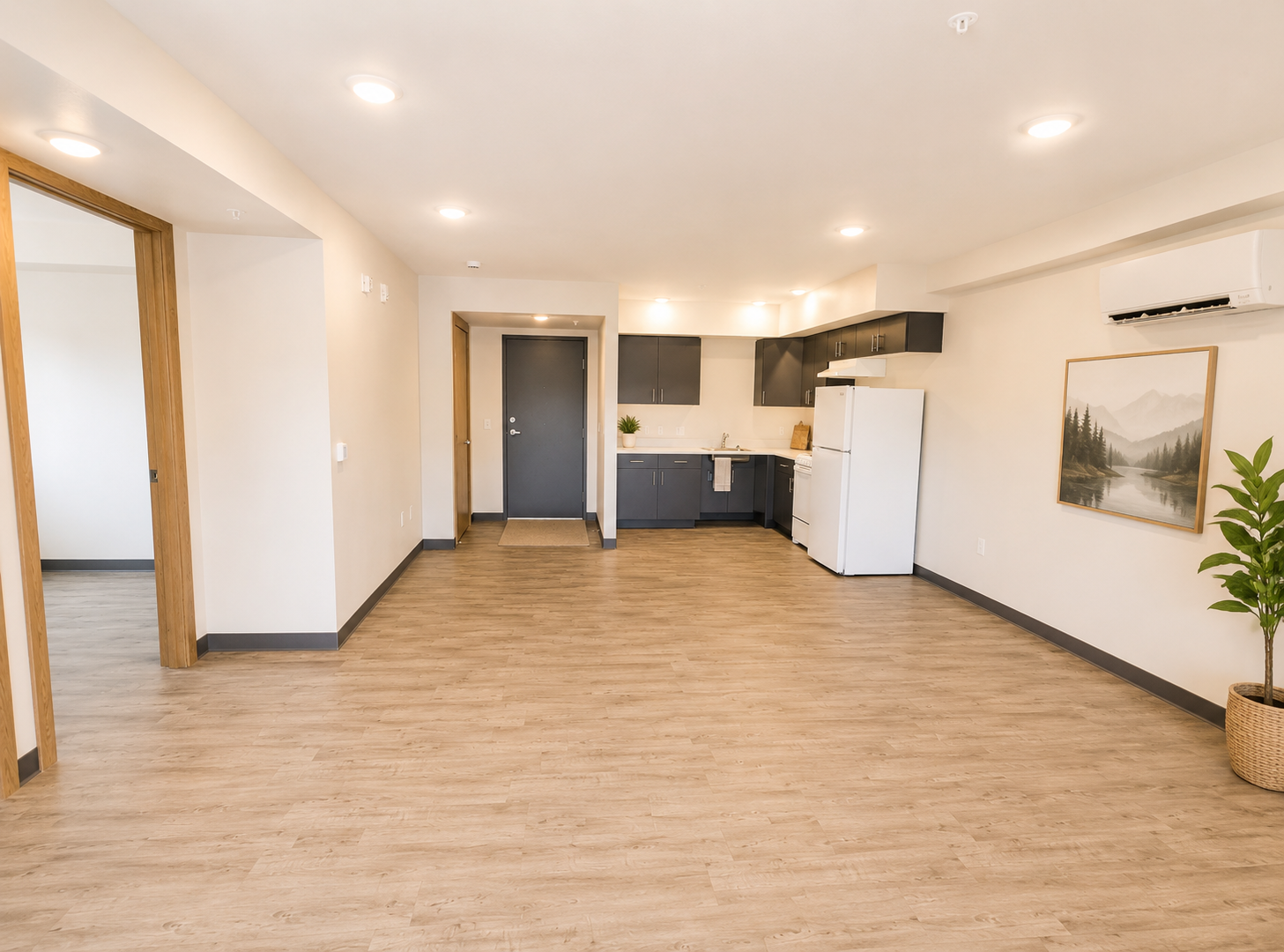 Spacious interior of an apartment featuring a modern kitchen with appliances, light hardwood flooring, and a small potted plant in the corner. The room is bright with recessed lighting and includes a doorway leading to another area. A framed landscape picture is on the wall.