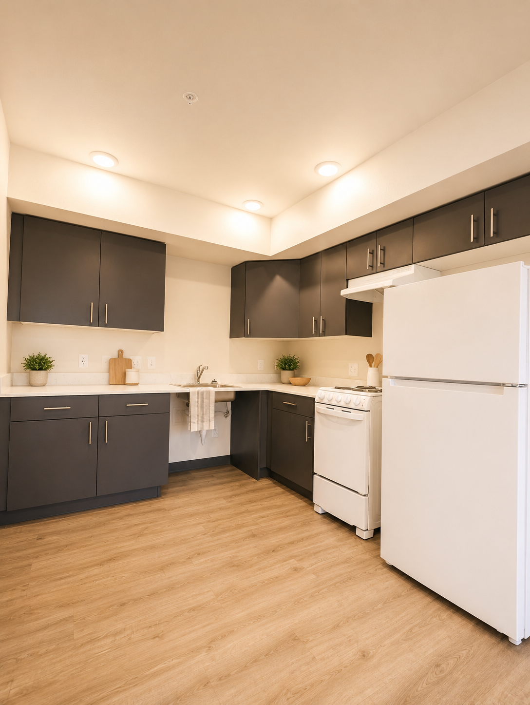 Modern kitchen featuring sleek dark cabinets, white countertops, a white stove, and a refrigerator. The space has a light wood floor and subtle green plants for decoration. Bright overhead lighting illuminates the room, creating a warm and inviting atmosphere.