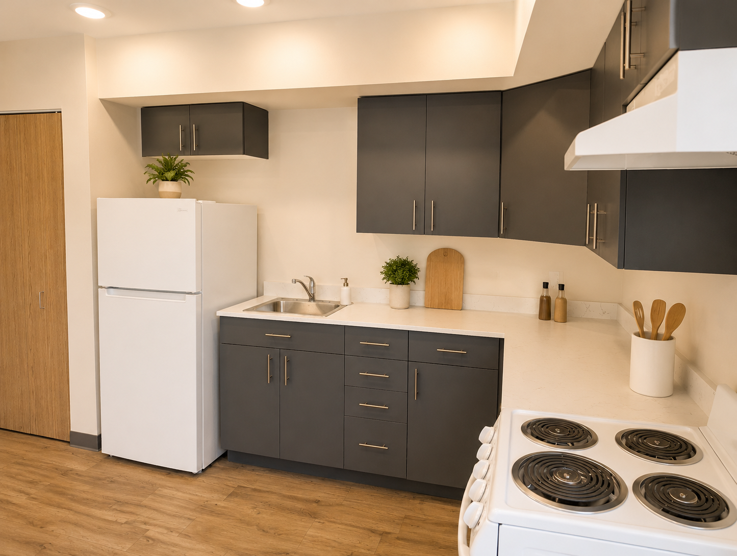 A modern kitchen featuring gray cabinetry, a white refrigerator, and a stainless steel sink. The countertop is light-colored, with a wooden cutting board and utensils displayed. The flooring is a warm wood tone, and there are plants on the shelves, creating a clean and inviting atmosphere.