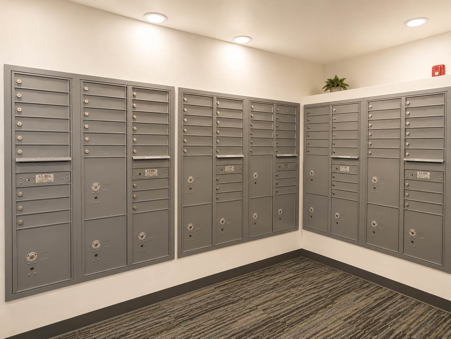 A view of a modern mailroom featuring multiple gray mailboxes arranged in a corner. The mailboxes have individual locking mechanisms and are mounted on the walls. Soft overhead lighting illuminates the area, highlighting the clean lines and contemporary design of the space, with a small plant in the corner.