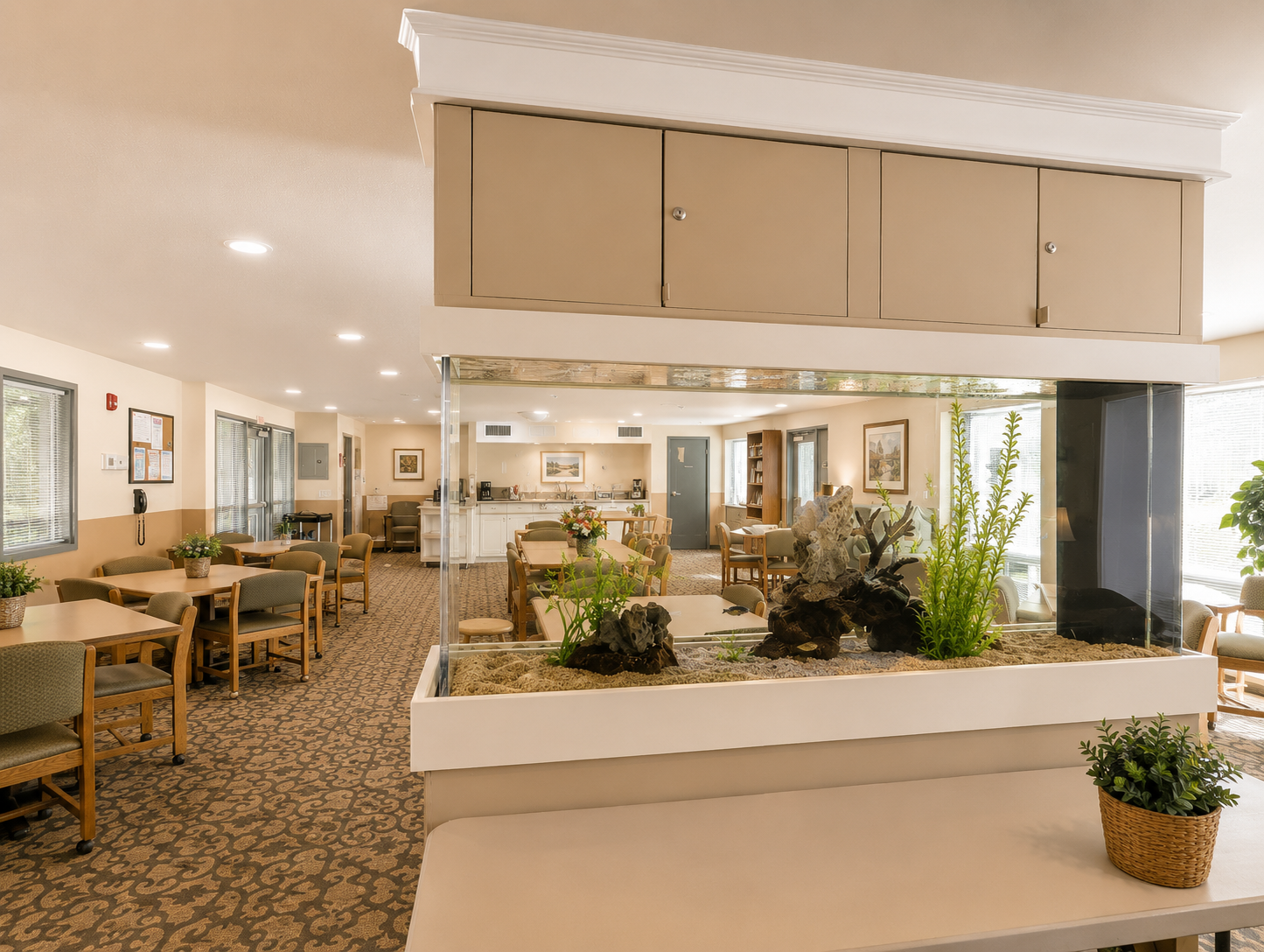 Interior of a welcoming common area featuring a large aquarium in the center, surrounded by light-colored furniture and carpet. Tables and chairs are arranged for dining or gathering, with plants adding greenery. Soft lighting creates a warm, inviting atmosphere.