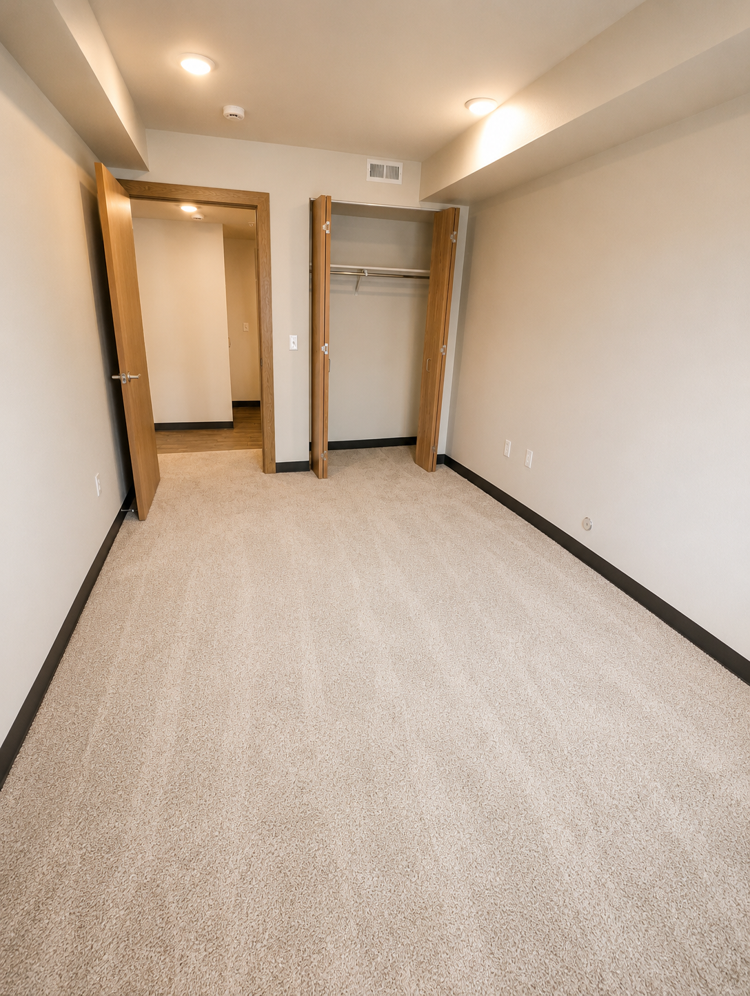 A spacious, empty room with light-colored carpet and neutral walls. Two doors are visible, leading to closets, and there are overhead lights illuminating the space. The room has a modern feel, with minimal furnishings, creating an open and clean atmosphere.