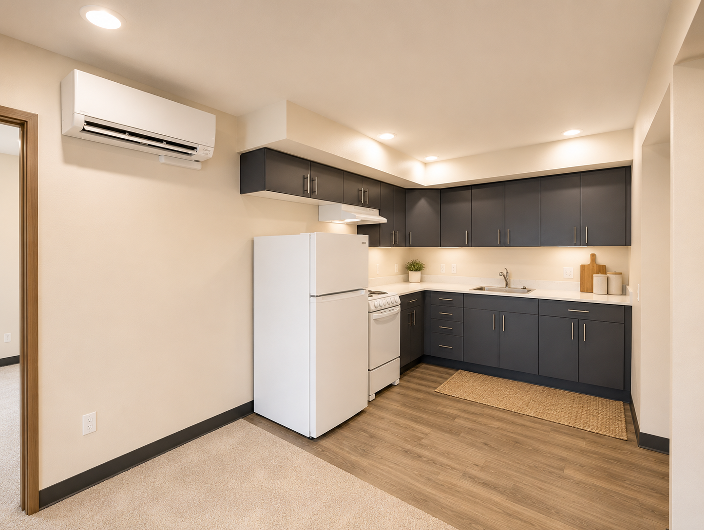 Modern kitchen featuring dark cabinetry, white appliances including a refrigerator and stove, a sink, and a small plant on the counter. The room has light-colored walls, recessed lighting, and a rug on the wood-like floor, creating a warm and inviting atmosphere.