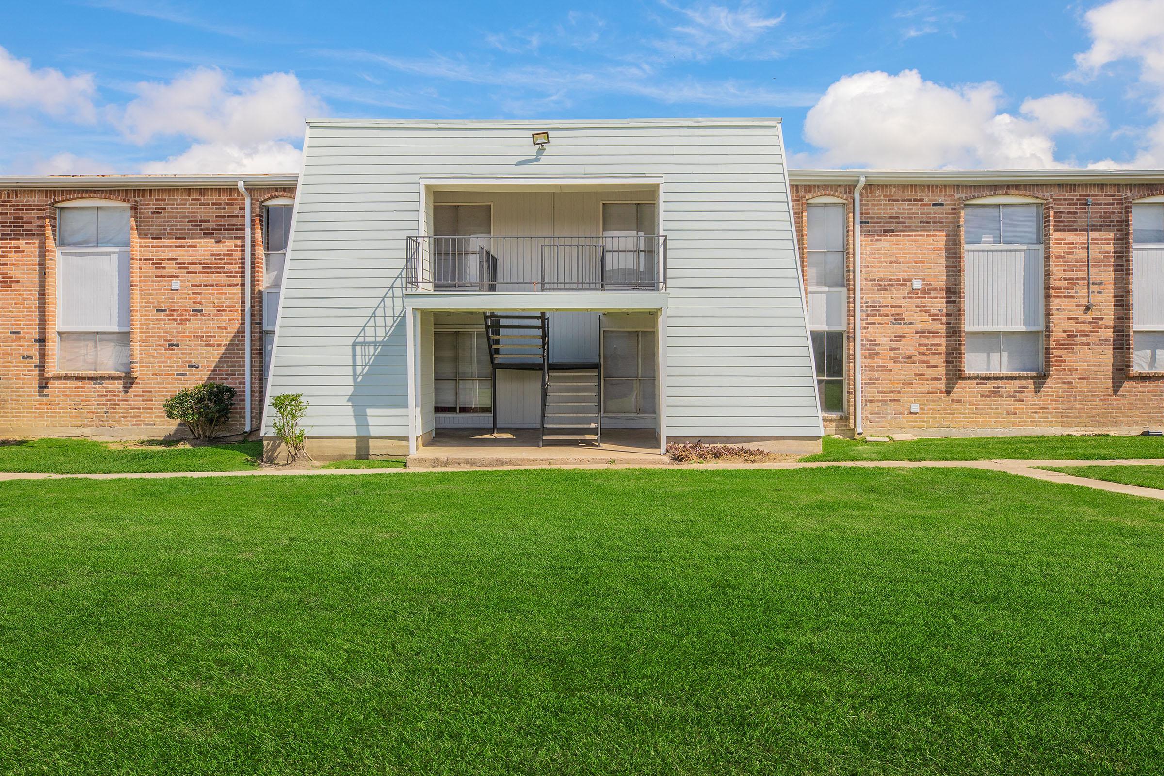 A two-story apartment building with a light blue facade and a distinctive triangular shape. The ground level features a balcony with a staircase leading up, surrounded by well-maintained green grass and landscaping. The sky is clear with a few clouds, creating a bright atmosphere.