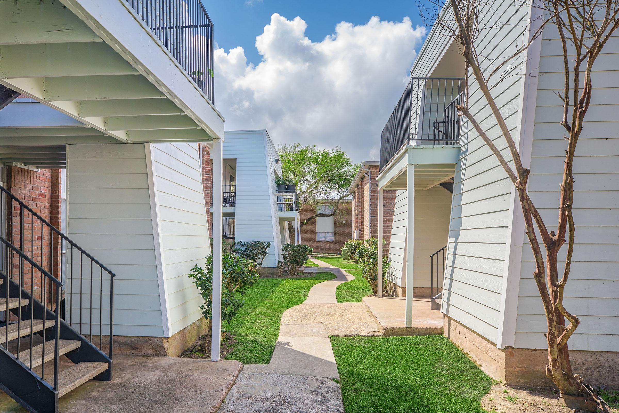 A serene scene of an apartment complex featuring two-story buildings with white siding. A well-maintained walkway winds through green lawns, bordered by shrubs. Soft clouds fill the bright blue sky, creating a calm atmosphere in the outdoor space.