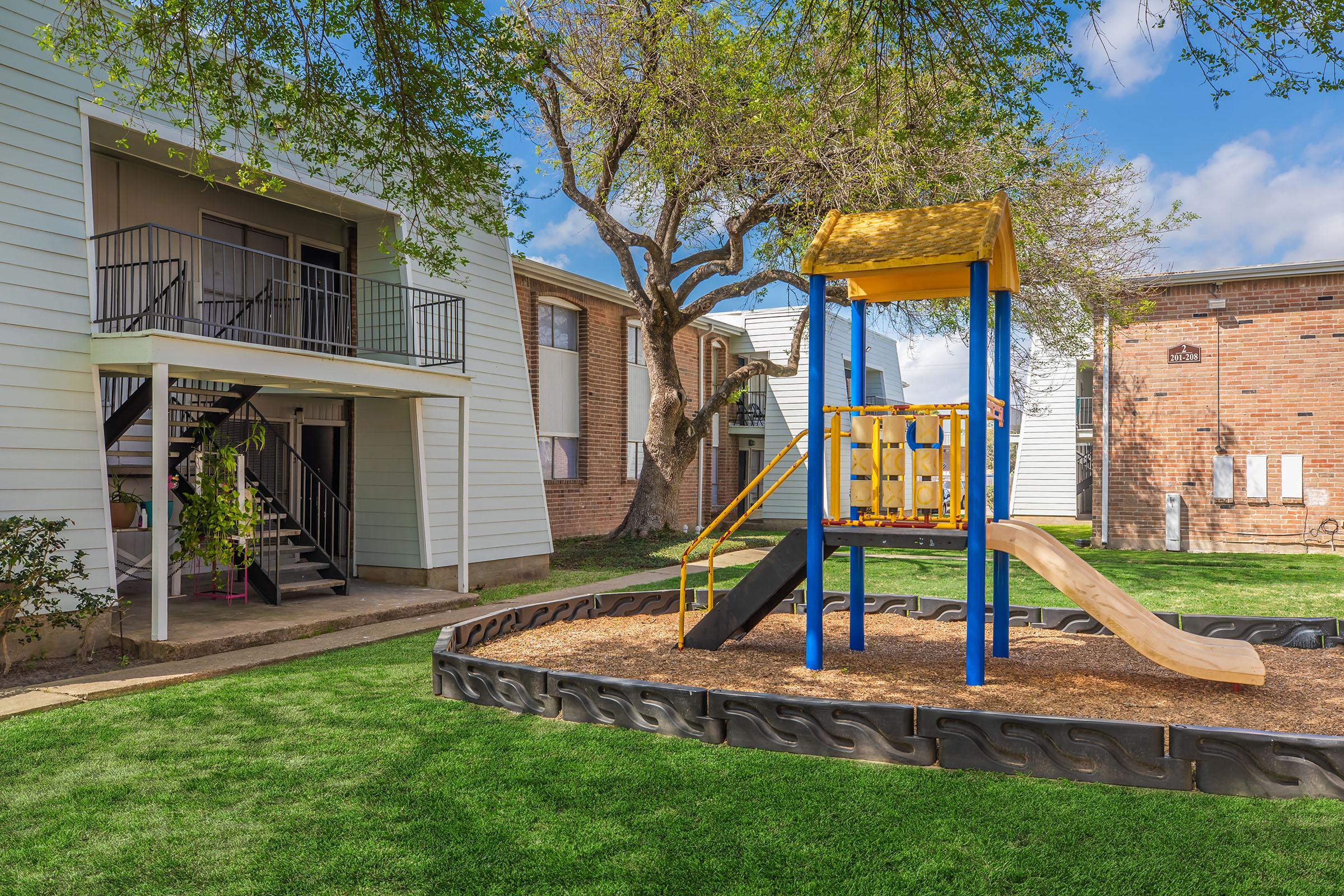 Playground with a yellow and blue slide structure set in a grassy area, surrounded by residential buildings. There's a tree providing shade, and an apartment building with a staircase and railing is visible in the background. The scene is bright and inviting, ideal for children to play.