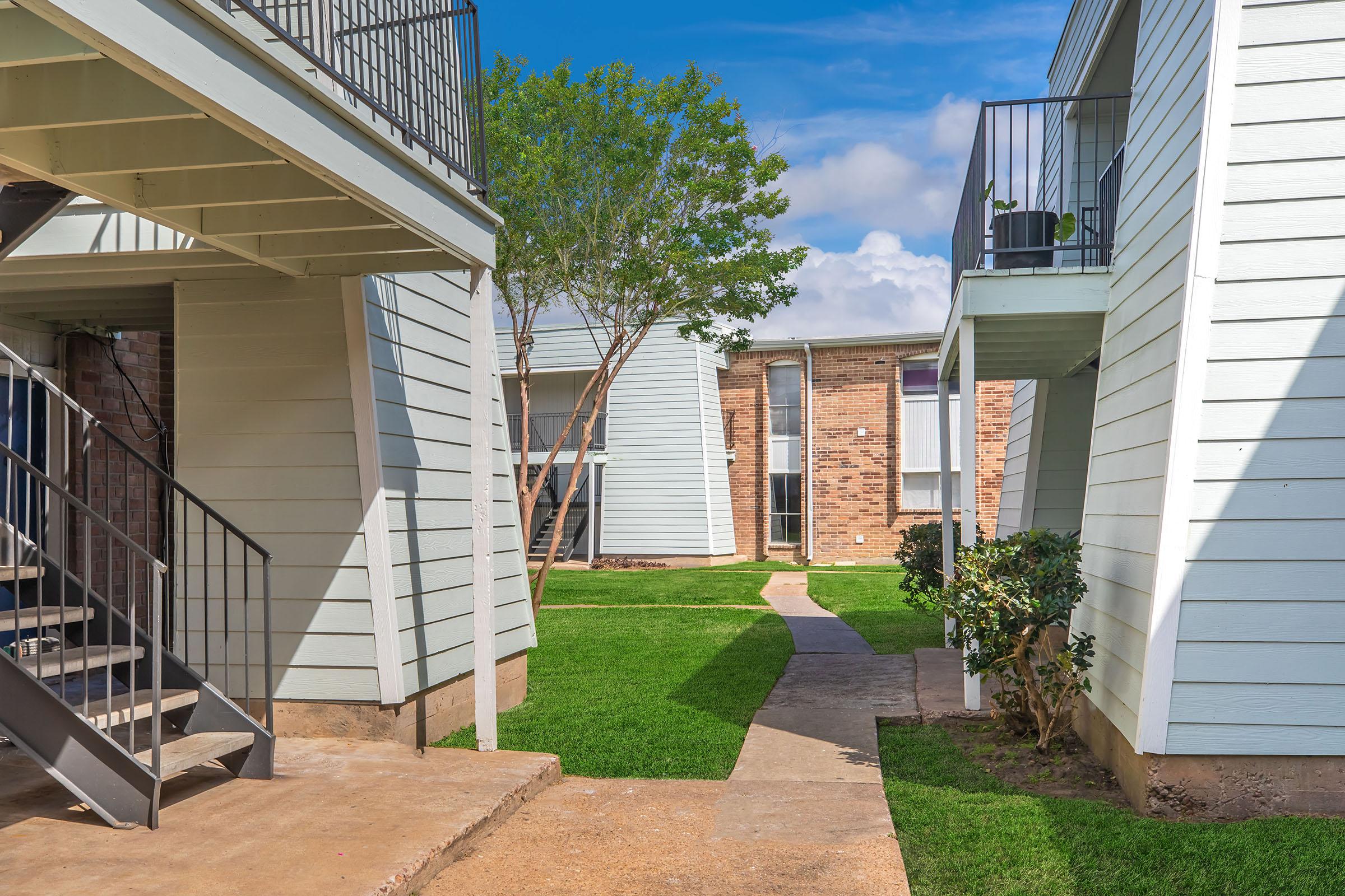 View of a residential complex with two sets of light-colored buildings on either side. A well-maintained pathway winds through a green lawn, leading to a brick building in the background. Trees are interspersed around the area, and there's a clear blue sky overhead, suggesting a sunny day.