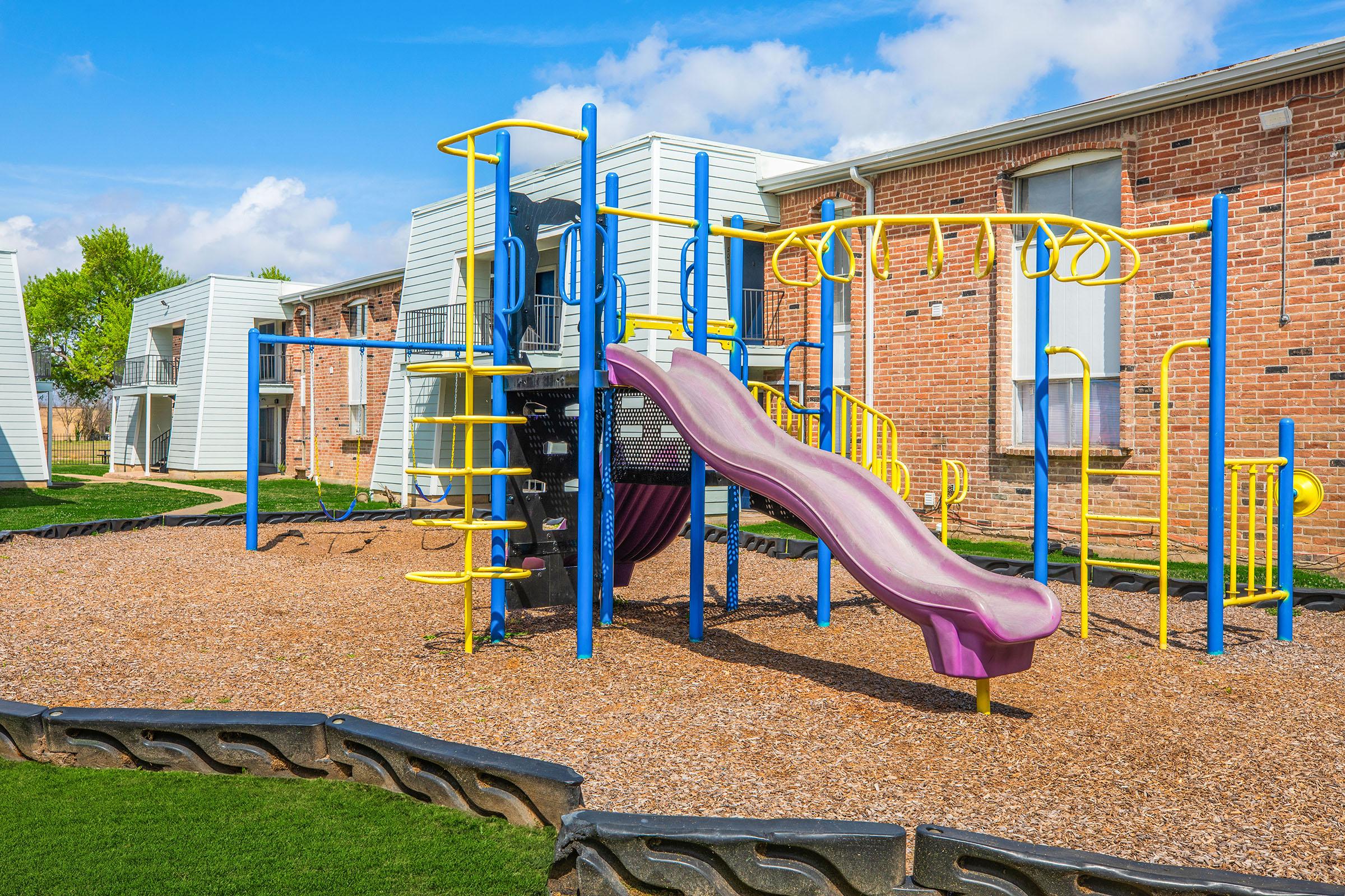 Colorful playground equipment with a purple slide, climbing structures, and yellow railings set on a sandy surface. Surrounding the playground are green lawns and residential buildings with brick exteriors, under a bright blue sky with scattered clouds.