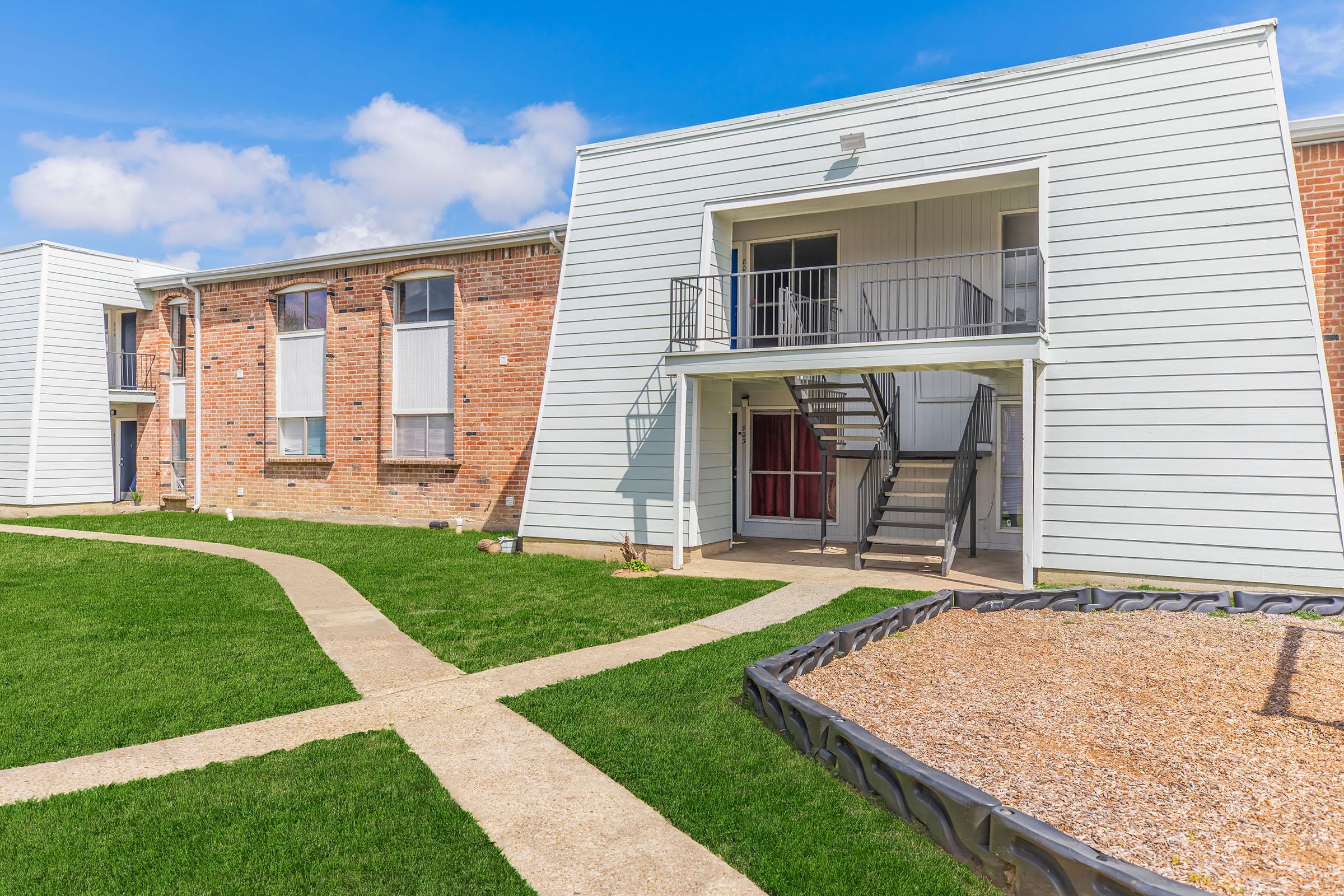 A well-maintained apartment complex with two buildings. The left building features exposed brick, while the right has a modern light-colored facade with balconies. Green grass surrounds a pathway leading to the entrance, and there's a gravel area nearby. The sky is partly cloudy, adding to the bright atmosphere.