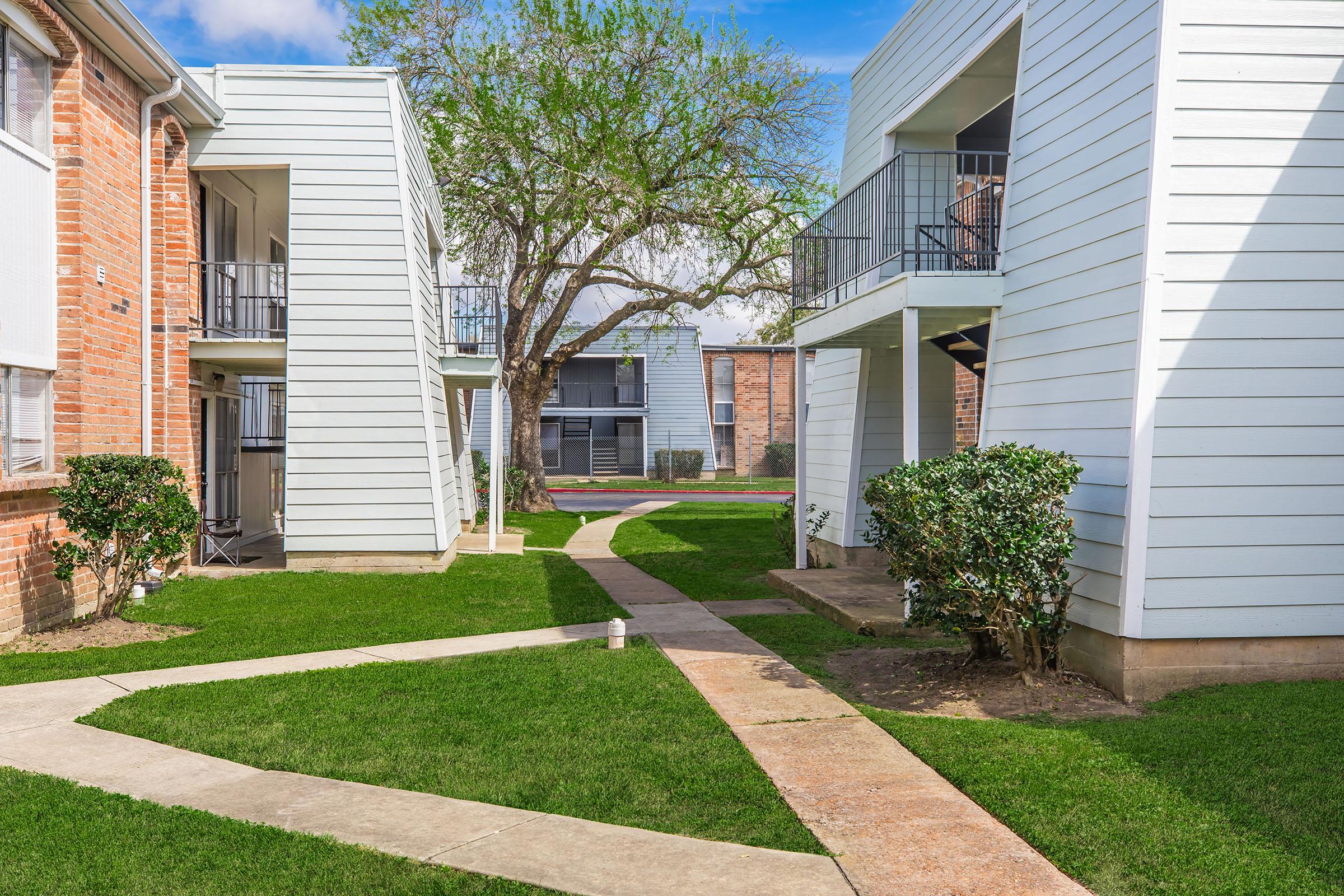 A view of an apartment complex featuring white and brick buildings with balconies, a landscaped green lawn, and a large tree. Pathways connect the buildings, leading to a central area, under a blue sky with scattered clouds.