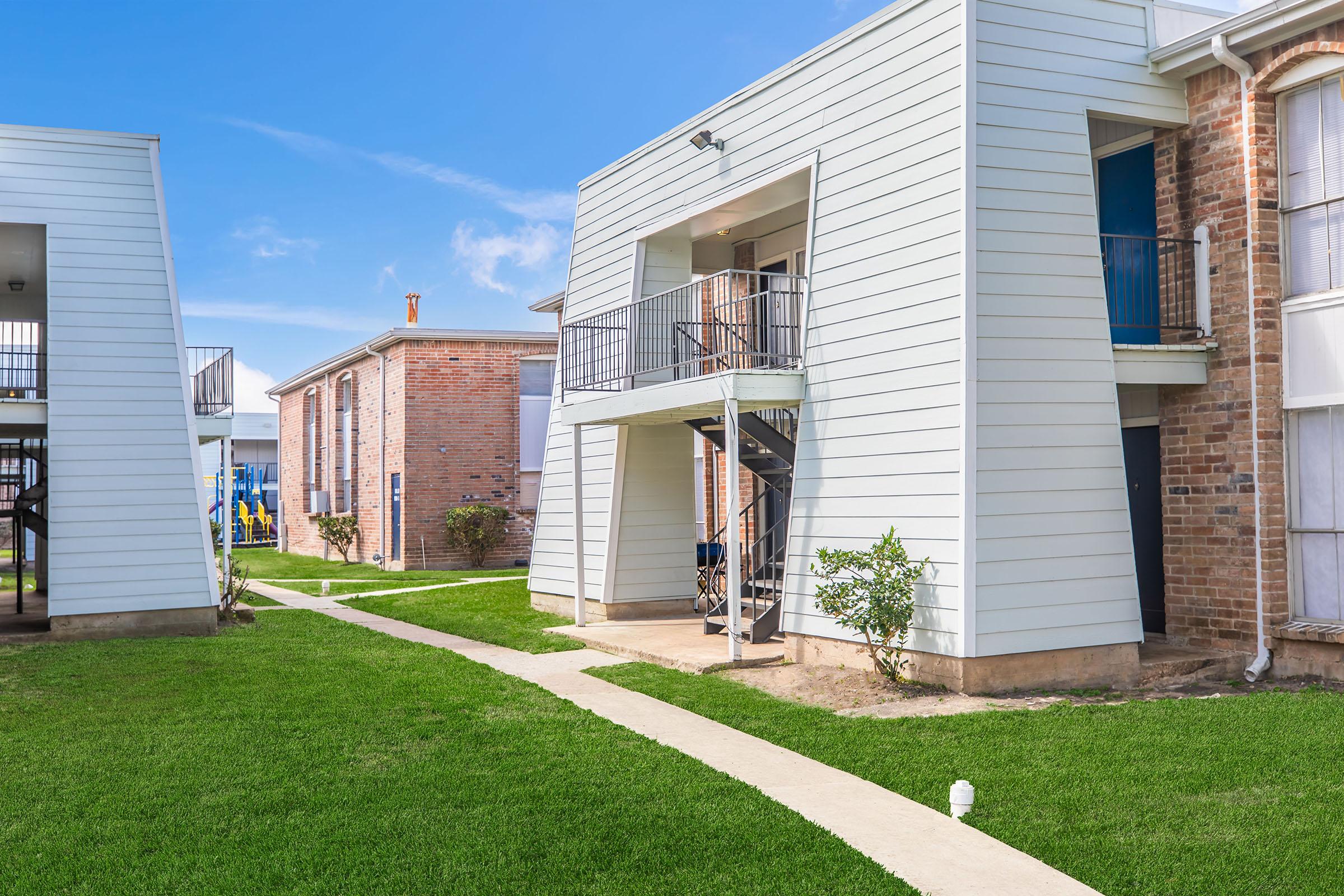 Two buildings in an apartment complex, one with a staircase leading to a balcony. The structures have a mix of white siding and brick exteriors. A paved walkway runs through well-maintained green grass between the buildings. Blue sky and sunshine contribute to a bright atmosphere.