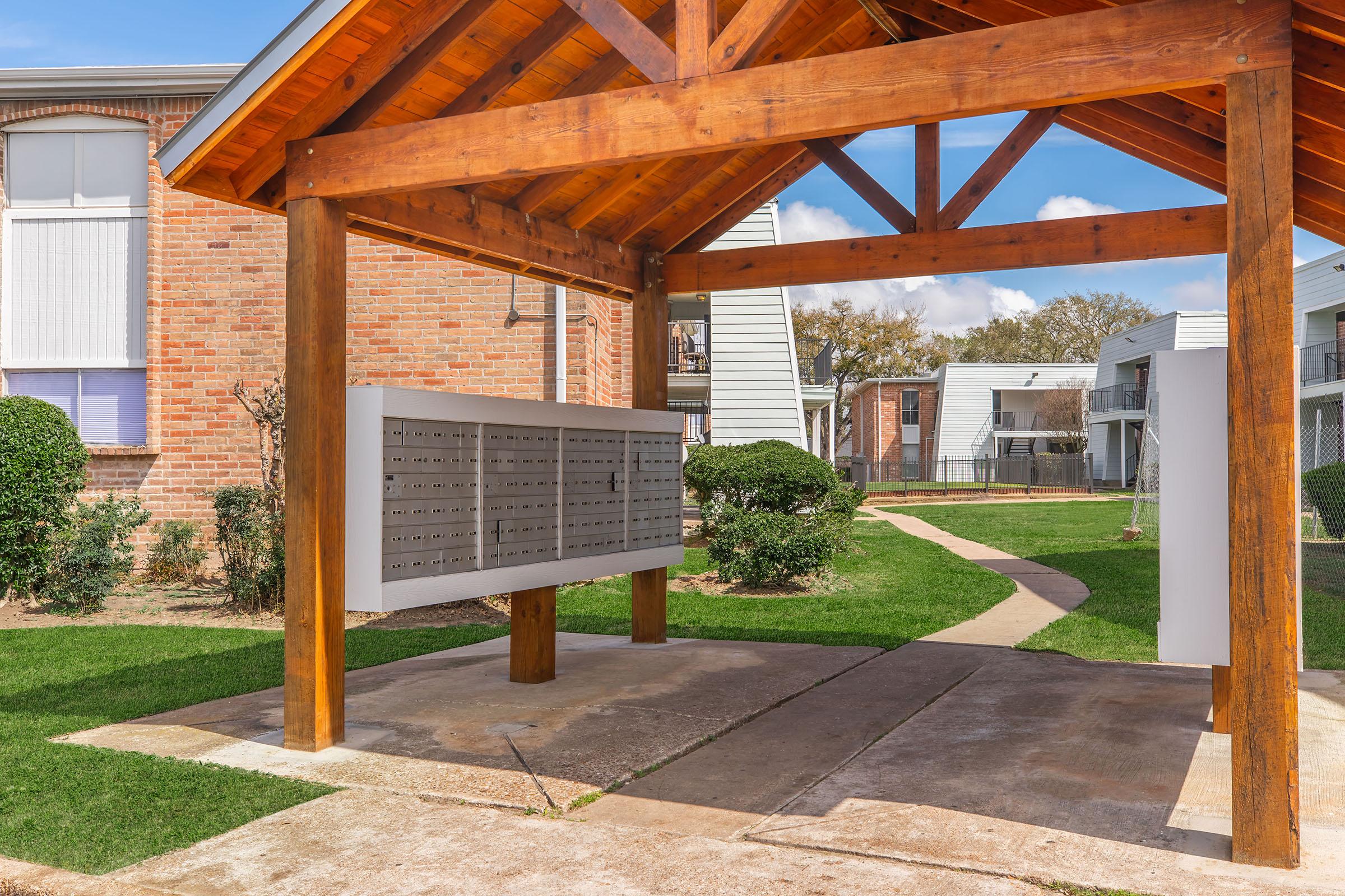 A covered mailbox area featuring a row of individual mailboxes on a concrete pad, surrounded by manicured grass and small hedges. There are residential buildings in the background, and a pathway leading through the green space. The structure has a wooden roof supported by posts.
