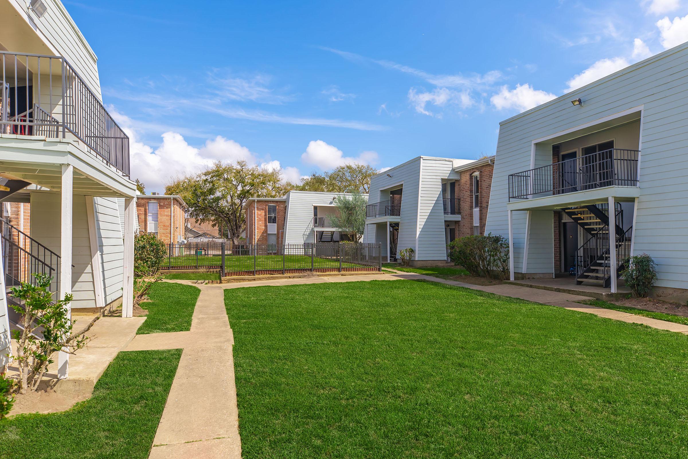 A landscaped area featuring several low-rise residential buildings with light-colored exteriors. There are manicured green lawns and concrete walkways connecting the buildings, with some trees and shrubs visible. The sky is partly cloudy, creating a bright and inviting atmosphere.
