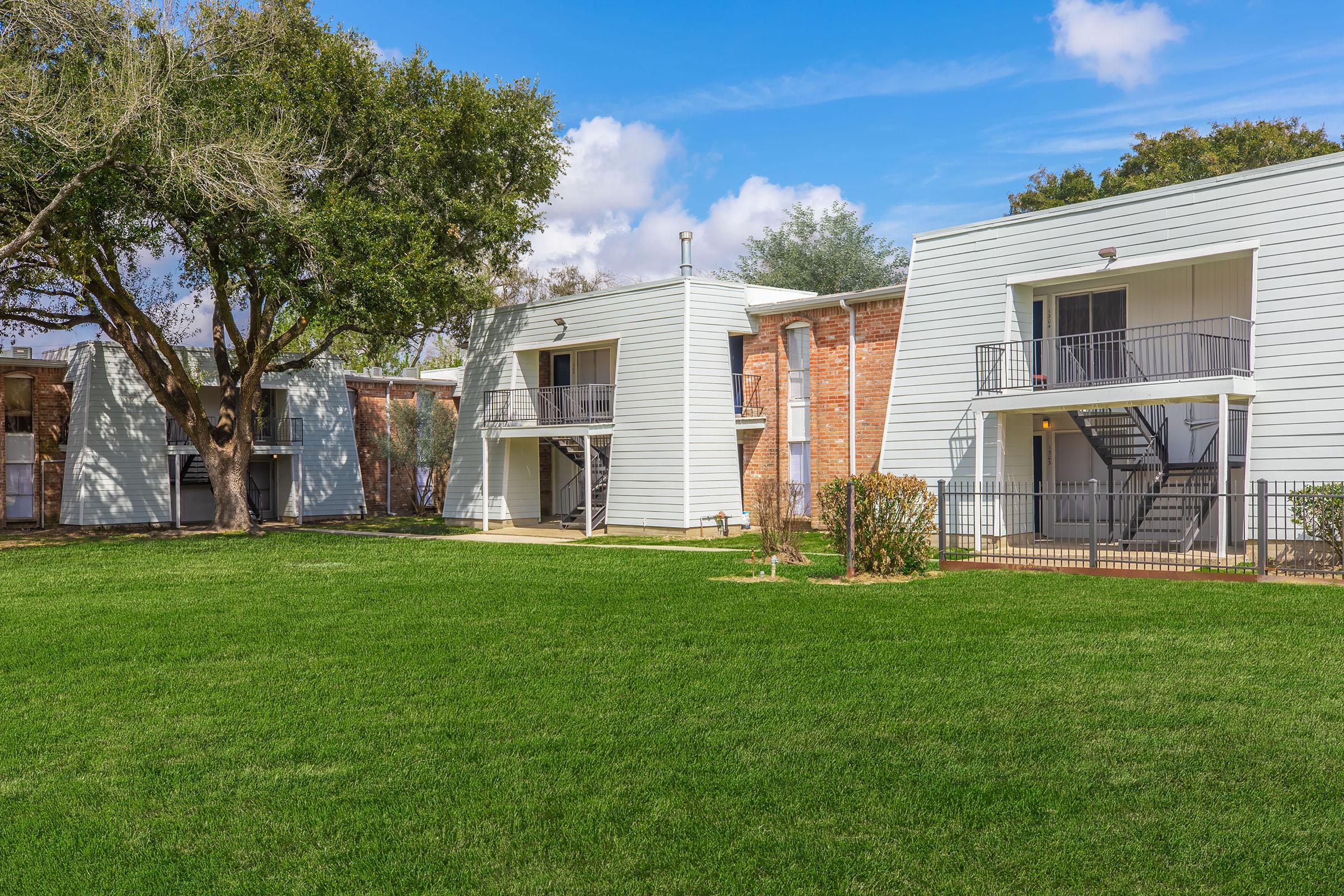 Two-story residential buildings with light-colored siding, surrounded by lush green grass. A large tree is visible to the left, and there are stairs leading up to second-floor balconies. The sky is clear with a few clouds, creating a bright and inviting atmosphere.