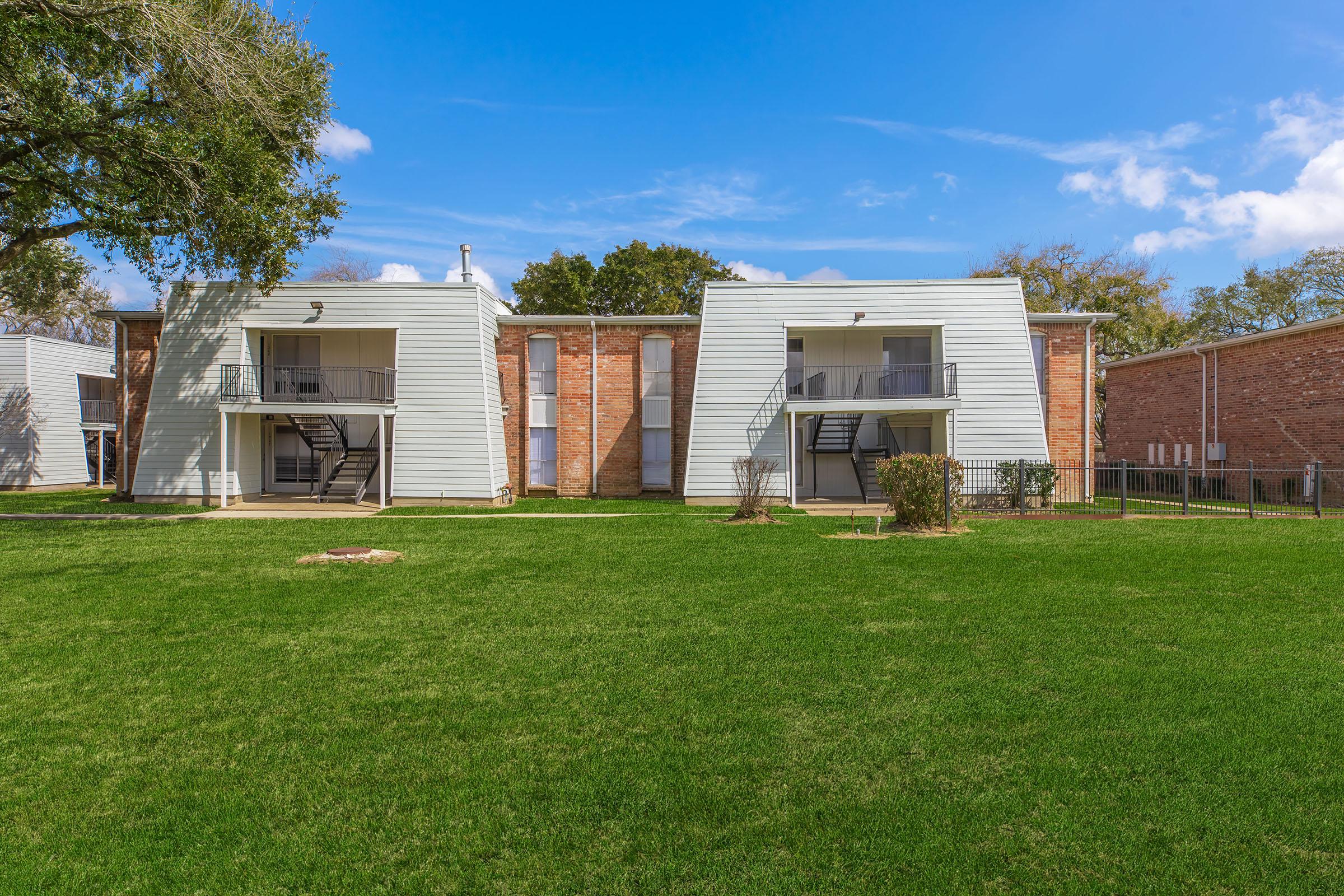 A bright, sunny day showcasing two adjacent apartment buildings with a green lawn in front. Each building features two stories with balconies and stairs. The sky is clear with a few clouds, and there are trees in the background, adding to the pleasant residential atmosphere.