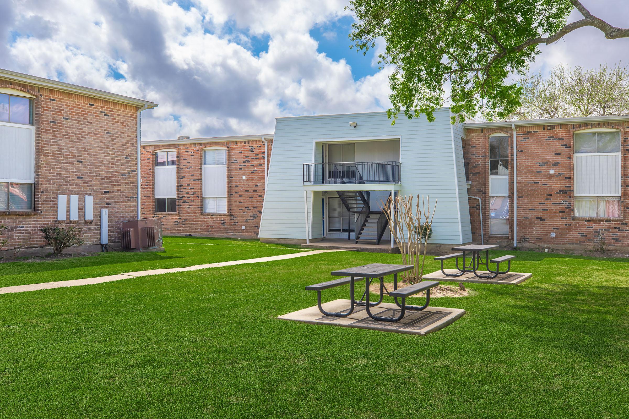 A view of an apartment complex with two brick buildings and a central grassy area. The foreground features picnic tables on a paved surface, while the background includes a staircase leading to a light-colored building facade. The scene is set under a partly cloudy sky with greenery around.