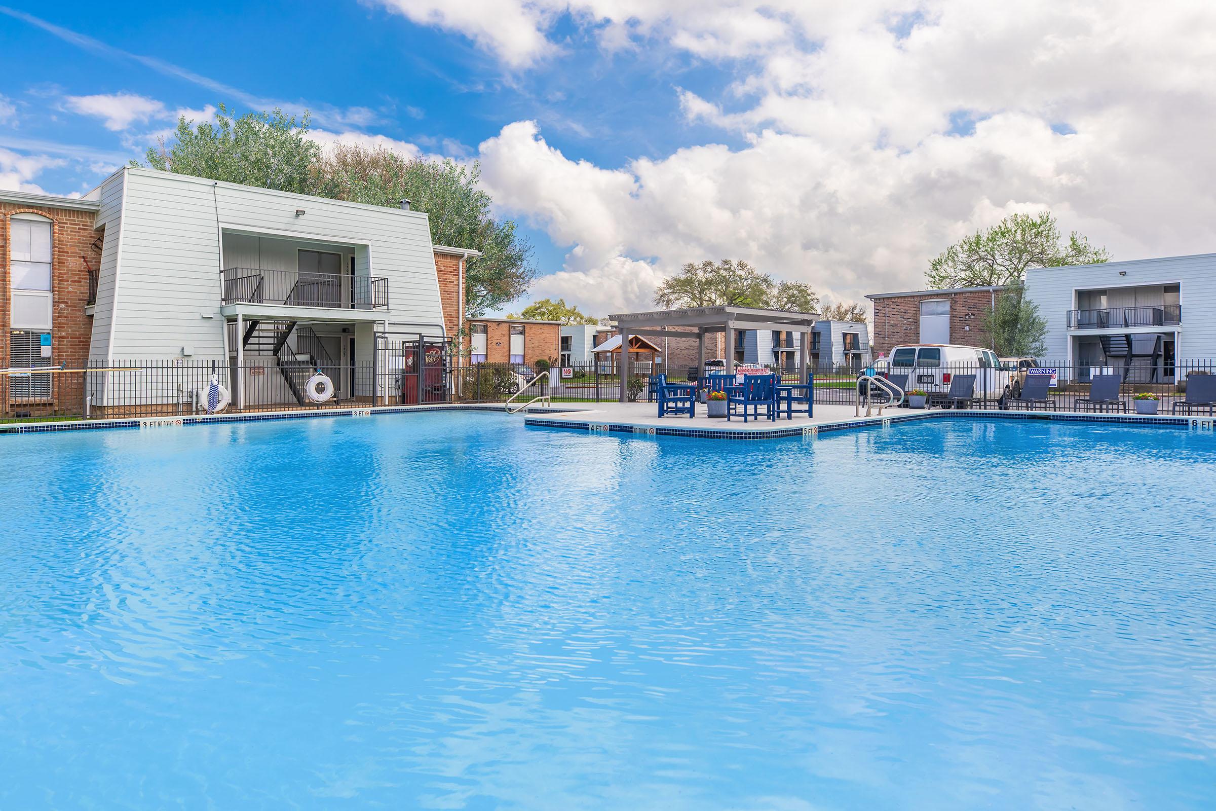 A clear blue swimming pool surrounded by residential buildings. The pool features lounge chairs and tables on a floating deck in the center. Lush greenery and a partly cloudy sky are visible in the background, creating a serene outdoor atmosphere.