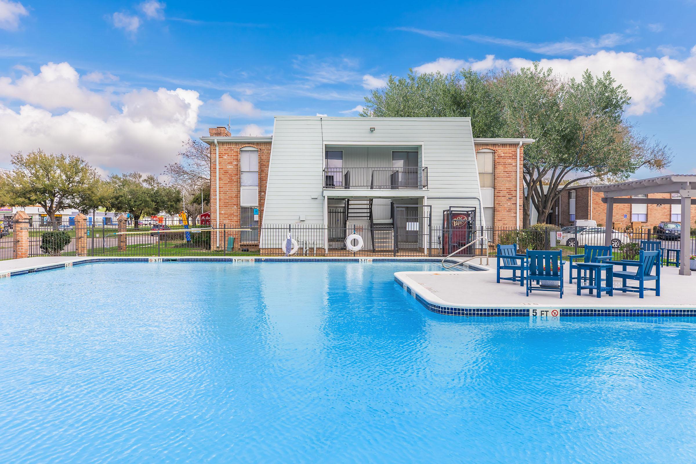 A clean, blue swimming pool surrounded by lounge chairs and a shaded seating area. In the background, a two-story building with a balcony is visible. The scene is set on a sunny day with partly cloudy skies and greenery nearby, creating a relaxing atmosphere.