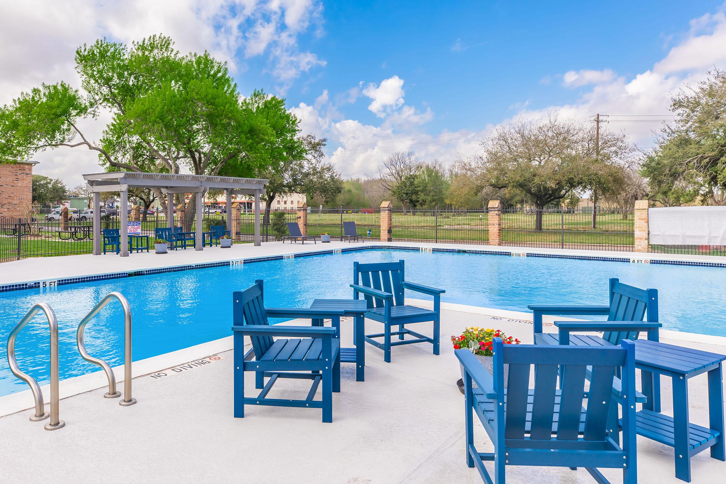A serene outdoor swimming pool surrounded by blue patio chairs and vibrant greenery. The pool area features a shaded seating structure and is bordered by a black fence, with a clear sky showcasing fluffy white clouds. Ideal for relaxation and leisure activities.