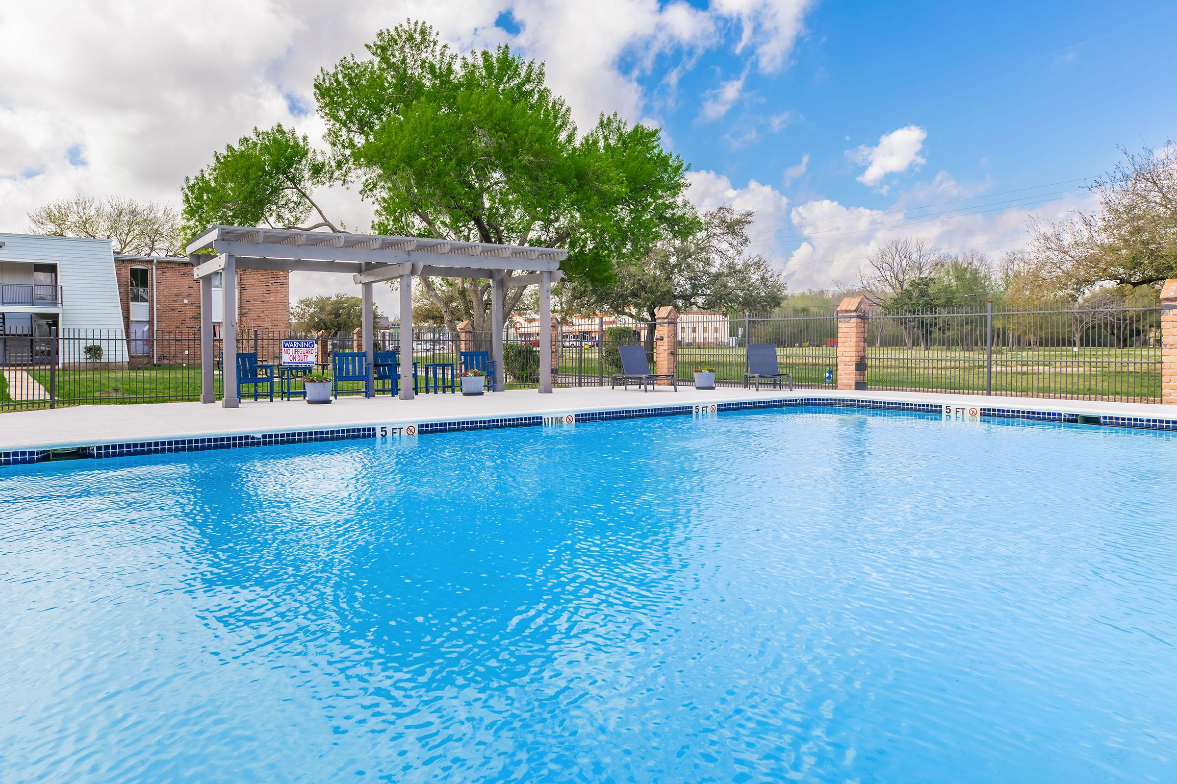 A clear blue swimming pool surrounded by a landscaped area, featuring a shaded pergola with seating. Lush green trees are visible in the background under a partly cloudy sky.