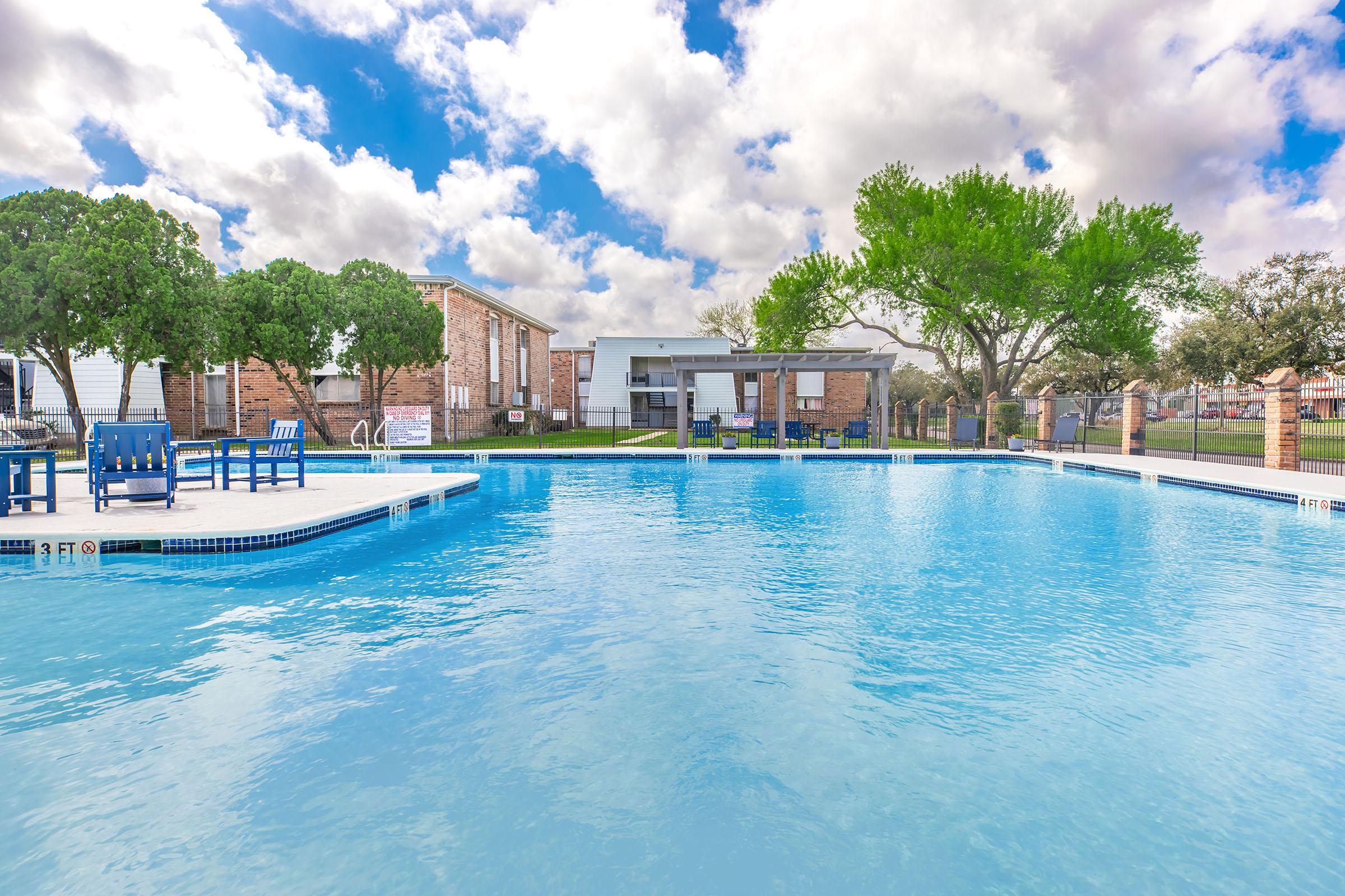 A clear blue swimming pool surrounded by lounge chairs under a partly cloudy sky. In the background, there is a modern building with large windows and greenery, including trees and grass. The scene is bright and inviting, perfect for relaxation and leisure.