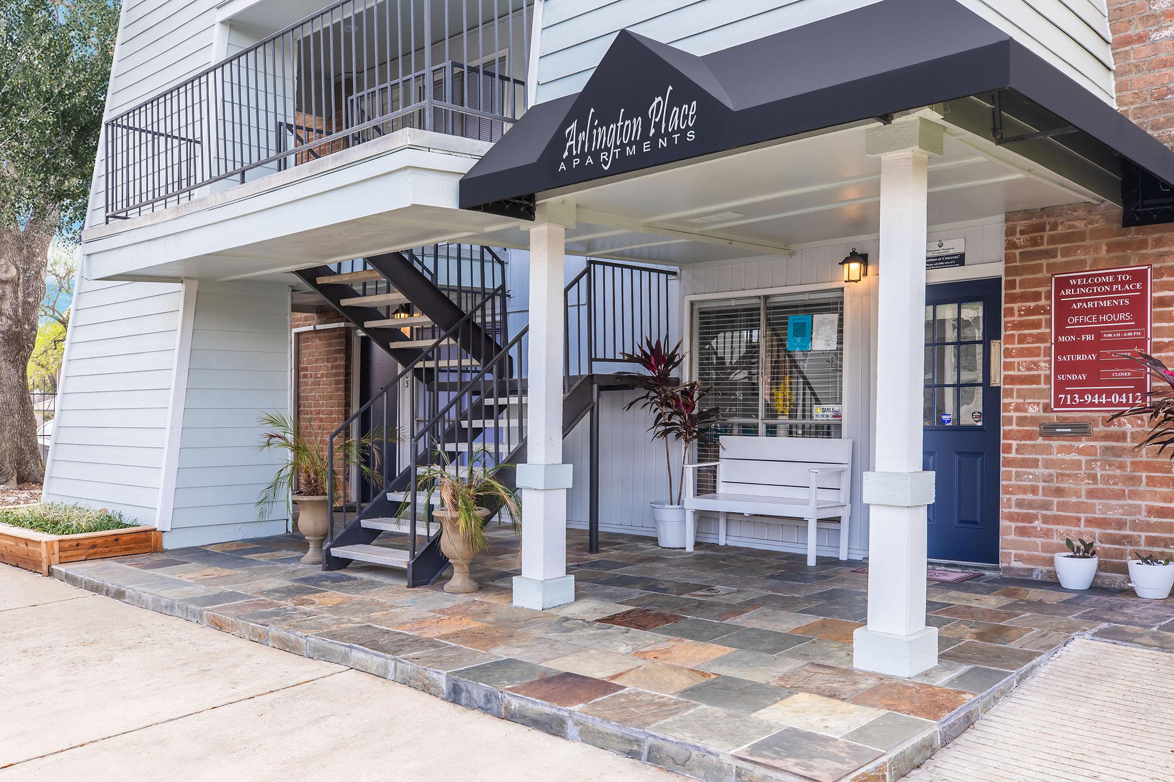 Entrance to Arlington Place Apartments featuring a welcoming porch with a bench, decorative plants, and a stairway leading to the upper floor. The building has a light blue exterior and brick accents, with a dark awning displaying the name of the apartments. Nearby is an informational sign with contact details.